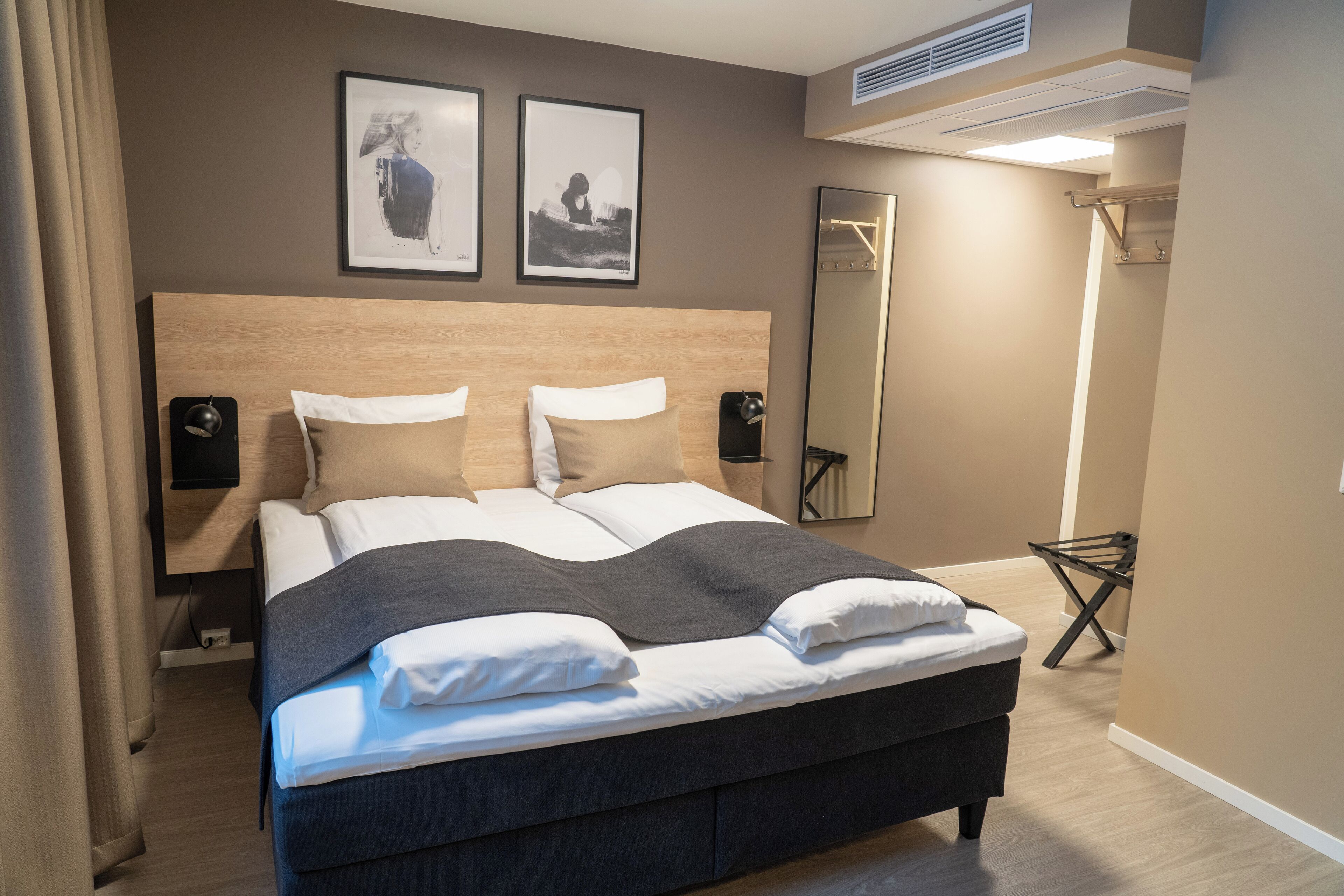 A neatly made bed with a dark headboard, two wall-mounted speakers, and two framed artworks above it.
