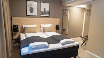 A neatly made bed with a dark headboard, two wall-mounted speakers, and two framed artworks above it.
