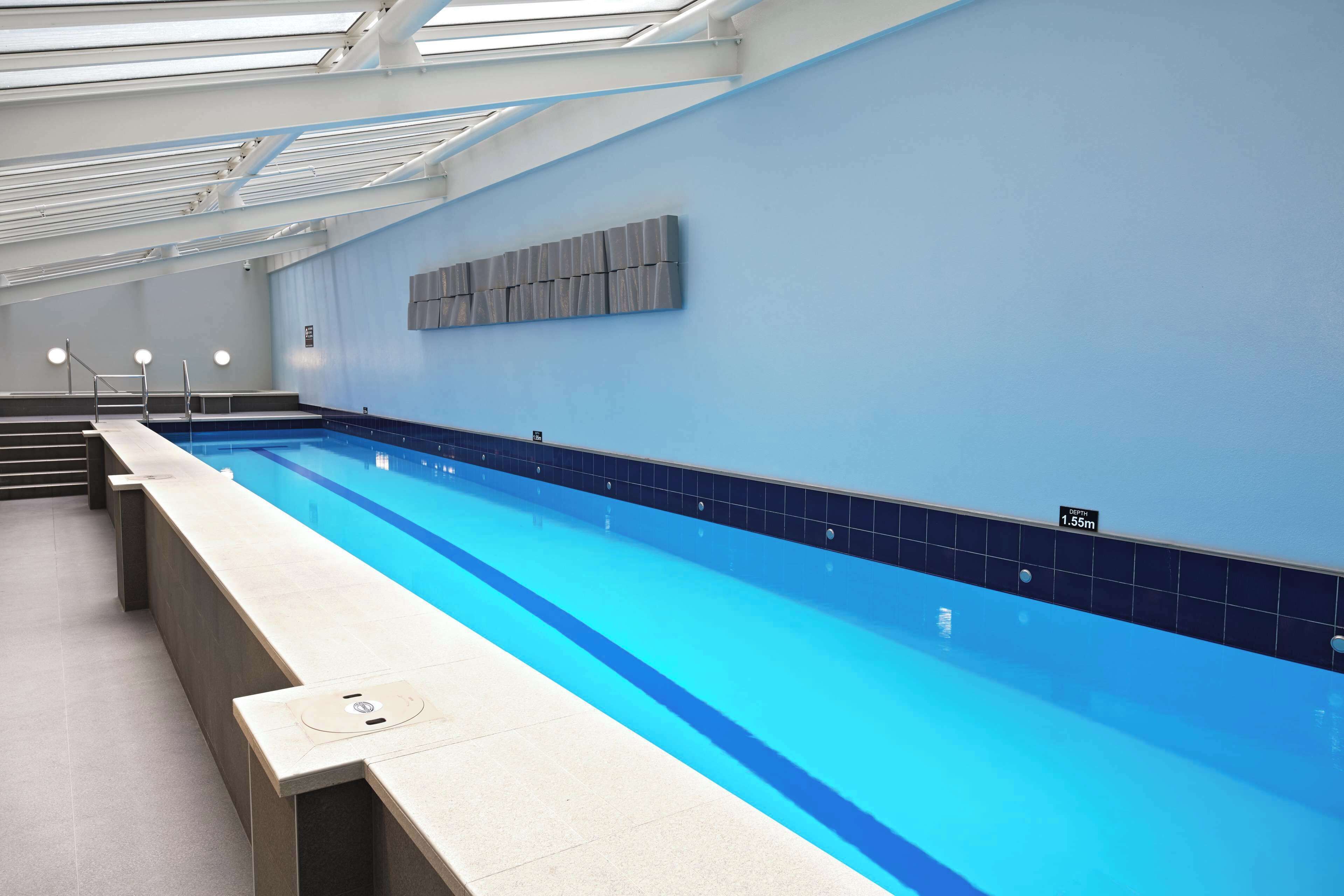 A long indoor swimming pool with a blue tiled edge, a depth sign, and a modern ceiling design.