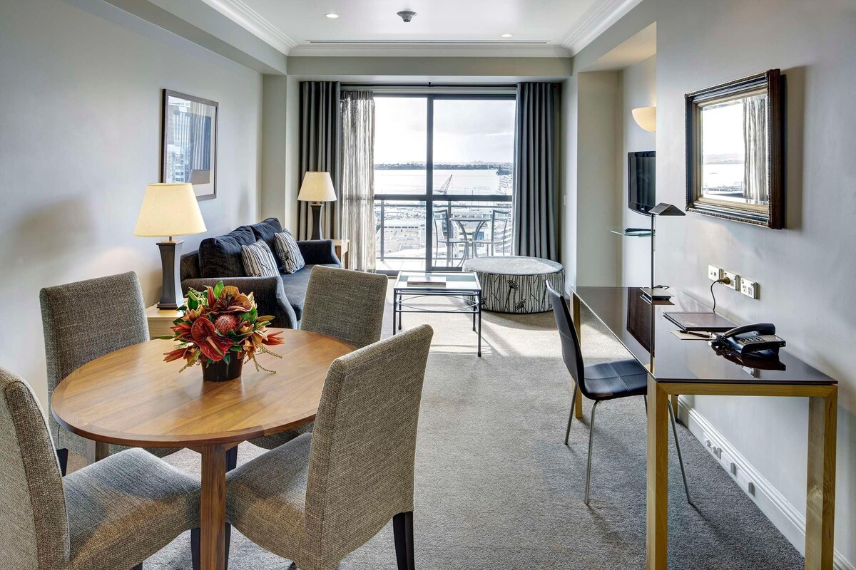 A hotel room with a dining area, a sofa, a TV, and a view of the city.