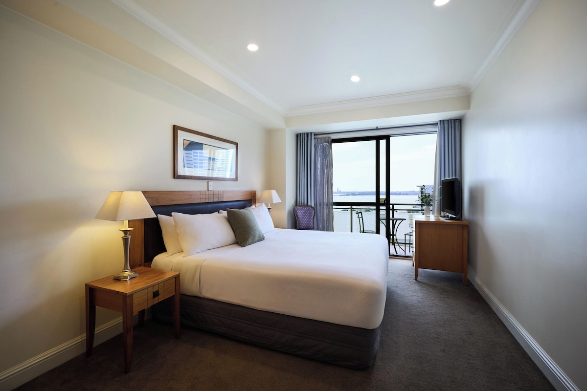Habitación de hotel con una cama grande, dos mesitas de noche, un televisor y un balcón con vistas.