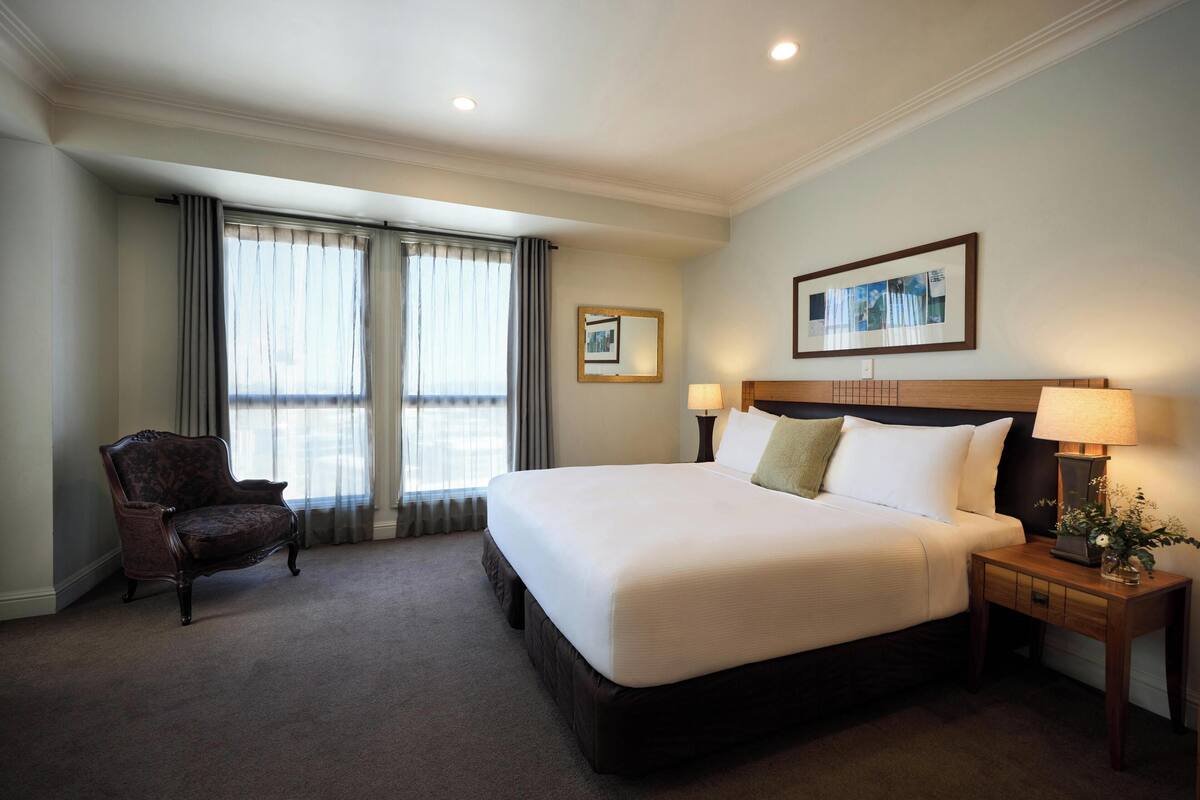 A hotel room with a large bed, two bedside lamps, a chair, a framed picture on the wall, and a window with curtains.