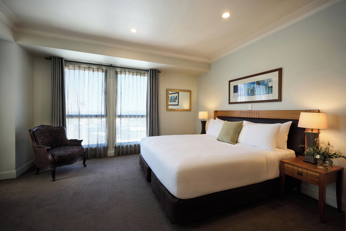 A hotel room with a large bed, two bedside lamps, a chair, a framed picture on the wall, and a window with curtains.