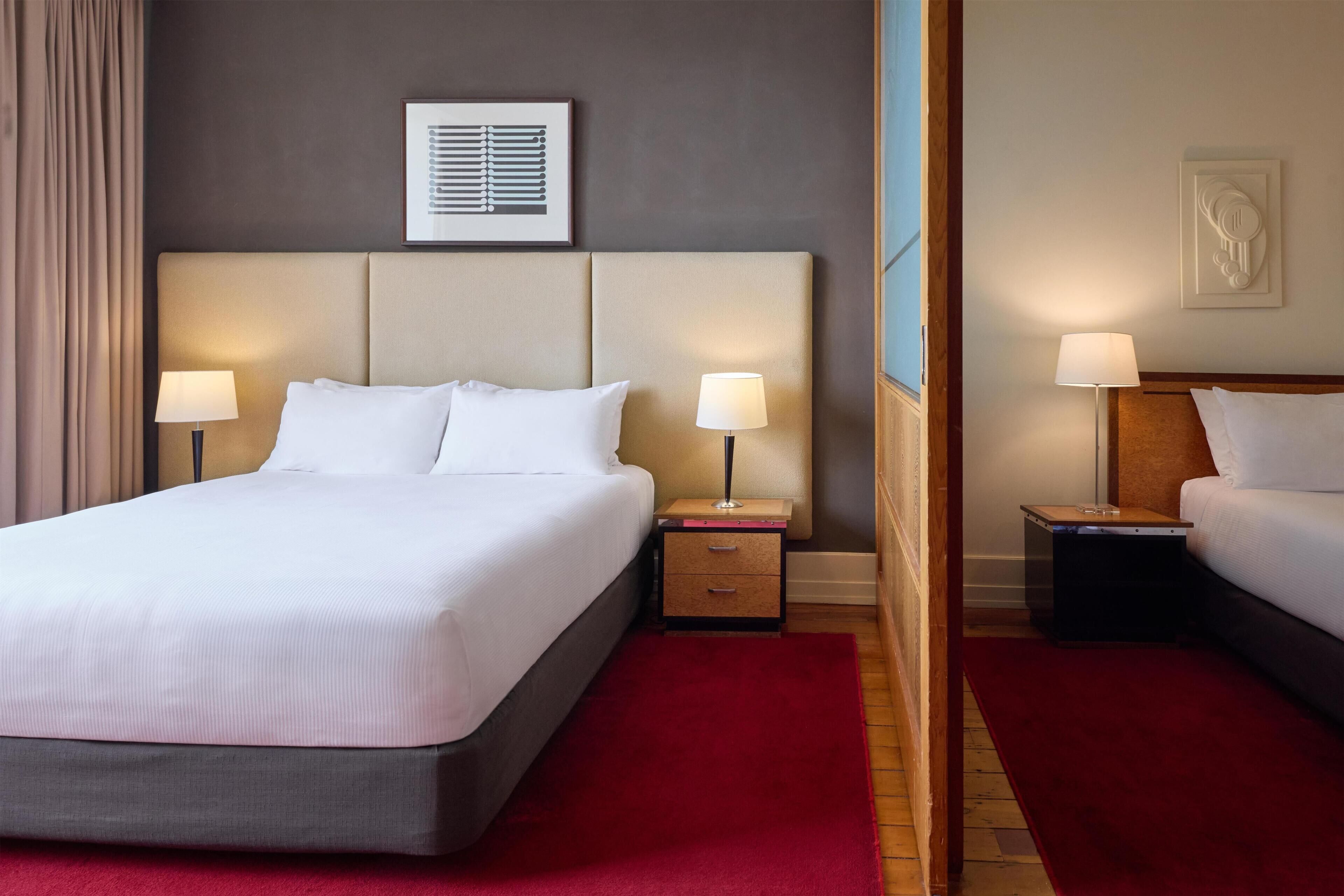Habitación de hotel con una cama grande, dos mesitas de noche, una alfombra roja y un cuadro en la pared.