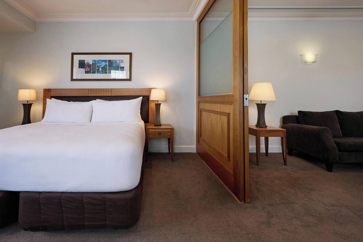 Une chambre d’hôtel avec un grand lit, deux tables de chevet avec des lampes, un canapé sombre, une armoire en bois et un tableau encadré au mur.