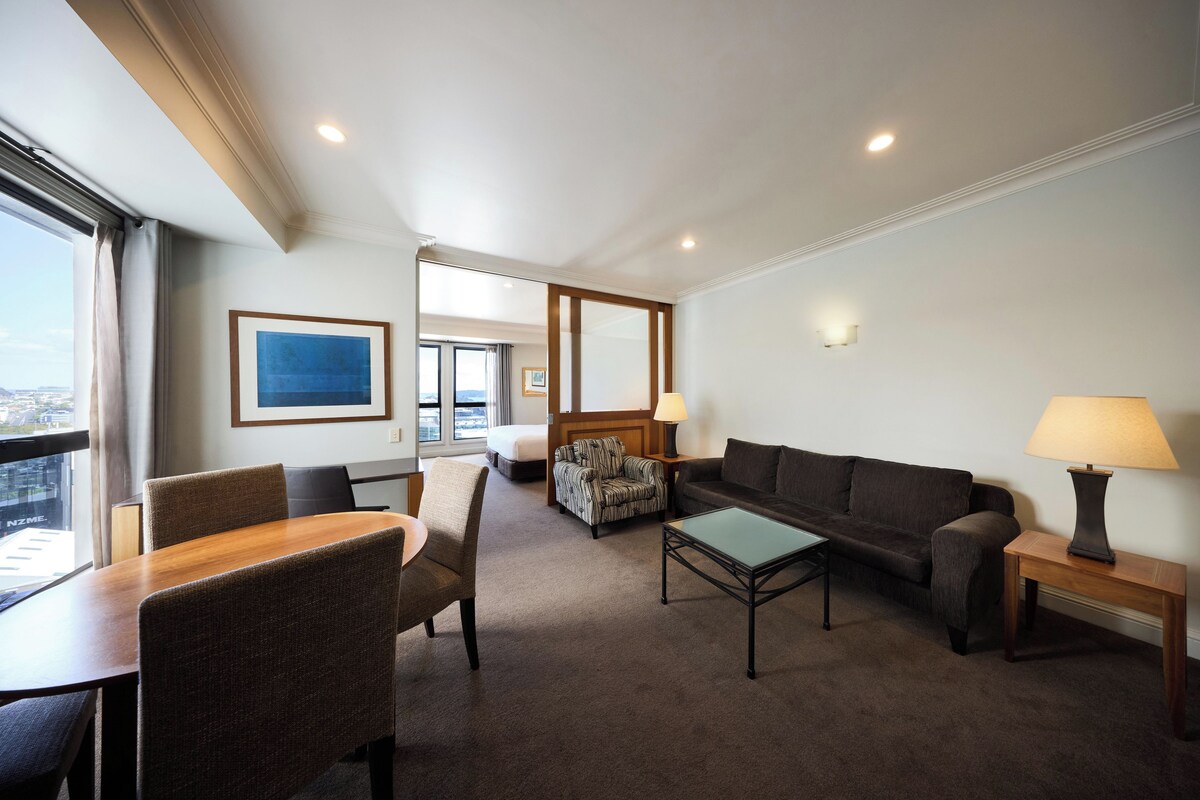 A hotel room with a dining table, two sofas, a patterned armchair, a lamp, and a framed picture on the wall.