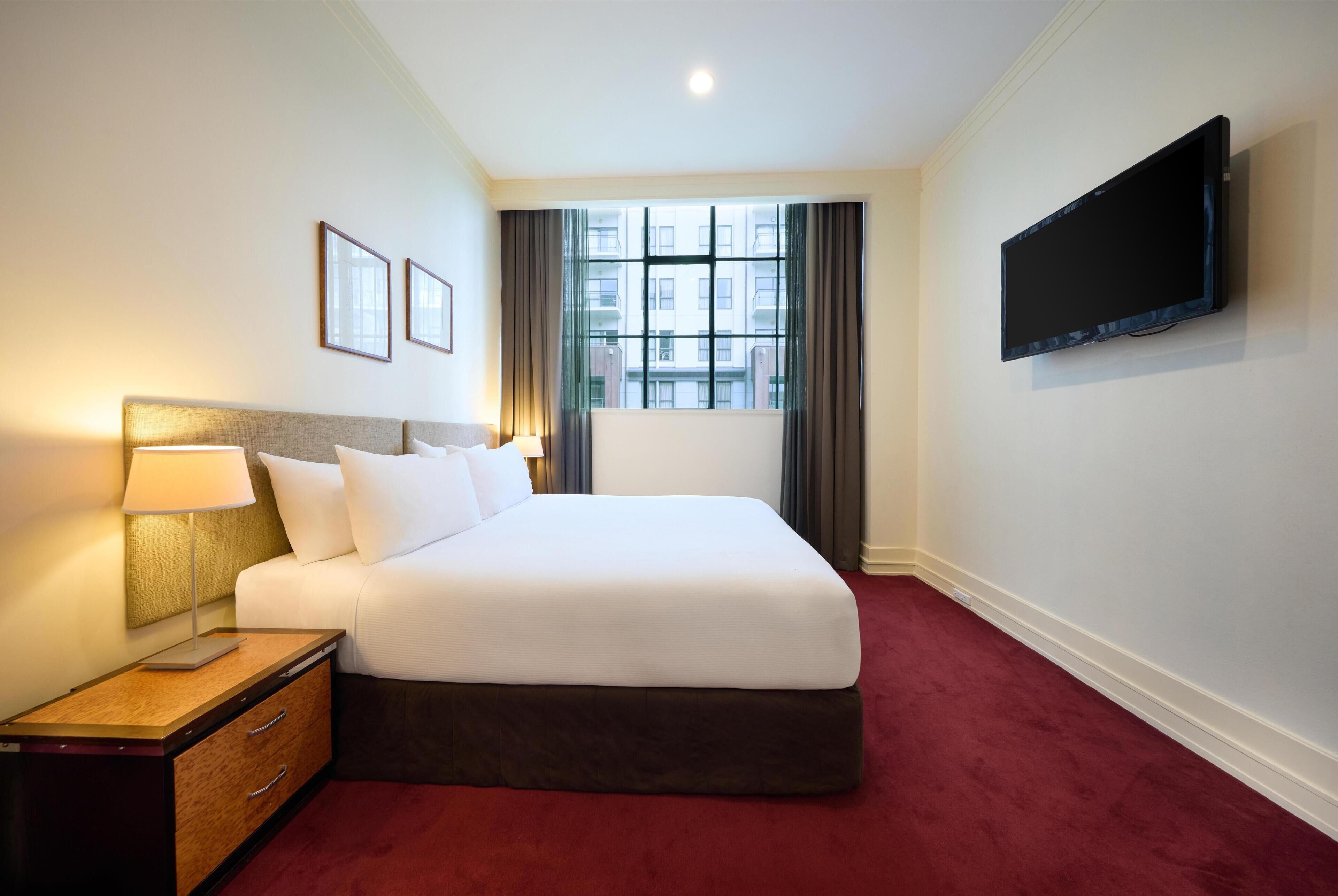 A hotel room with a bed, a flat-screen TV, a nightstand, a lamp, a window with curtains, and framed pictures on the wall.