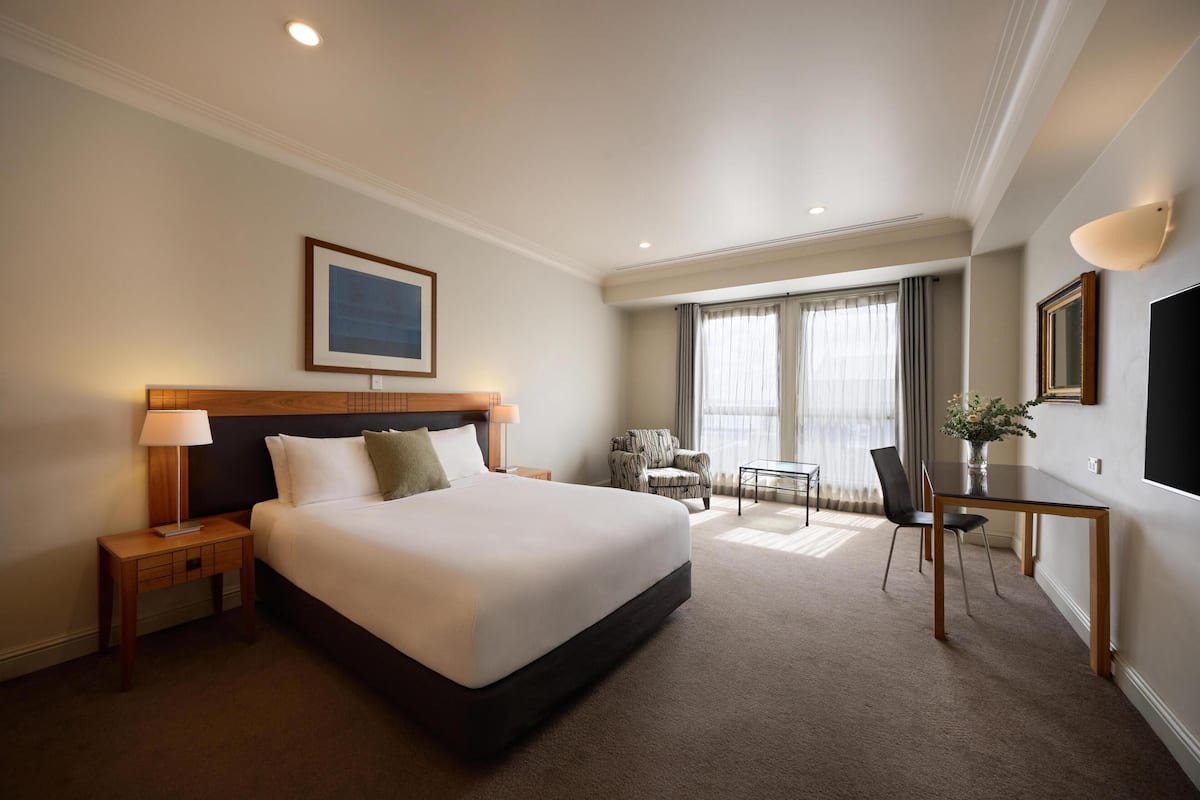 Une chambre d’hôtel comprenant un lit, une table de chevet, une lampe, une chaise, un bureau et une grande fenêtre avec des rideaux.