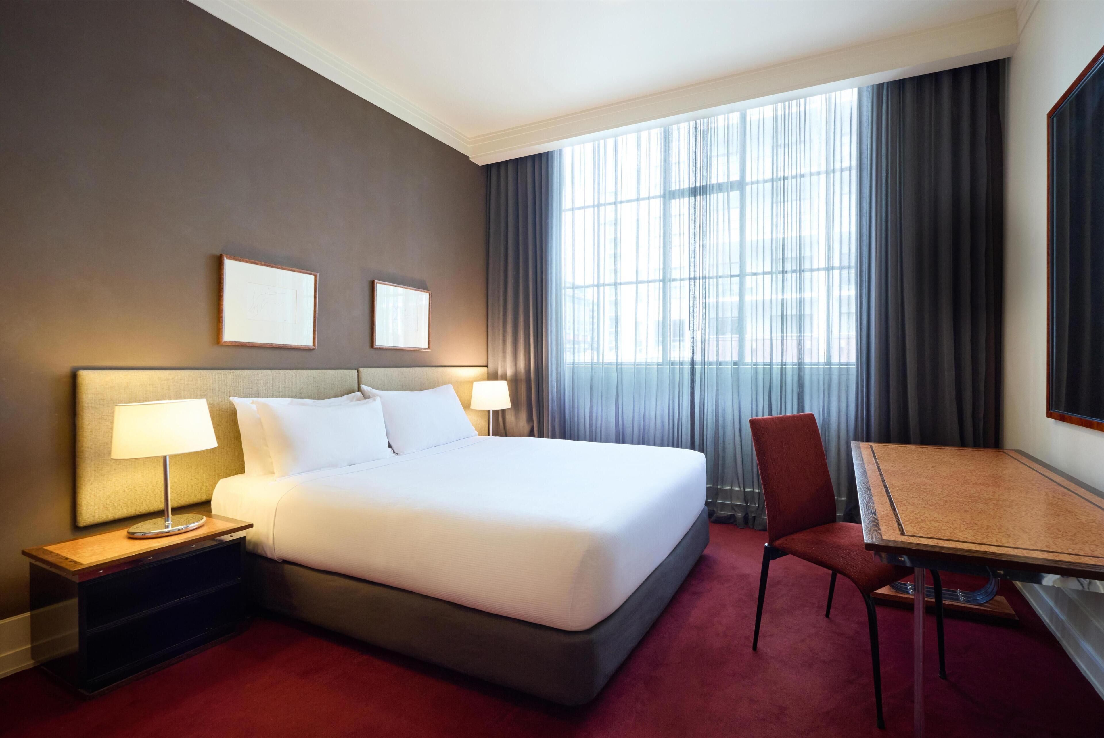 A hotel room with a large bed, a desk, a chair, two bedside lamps, and a window with curtains.