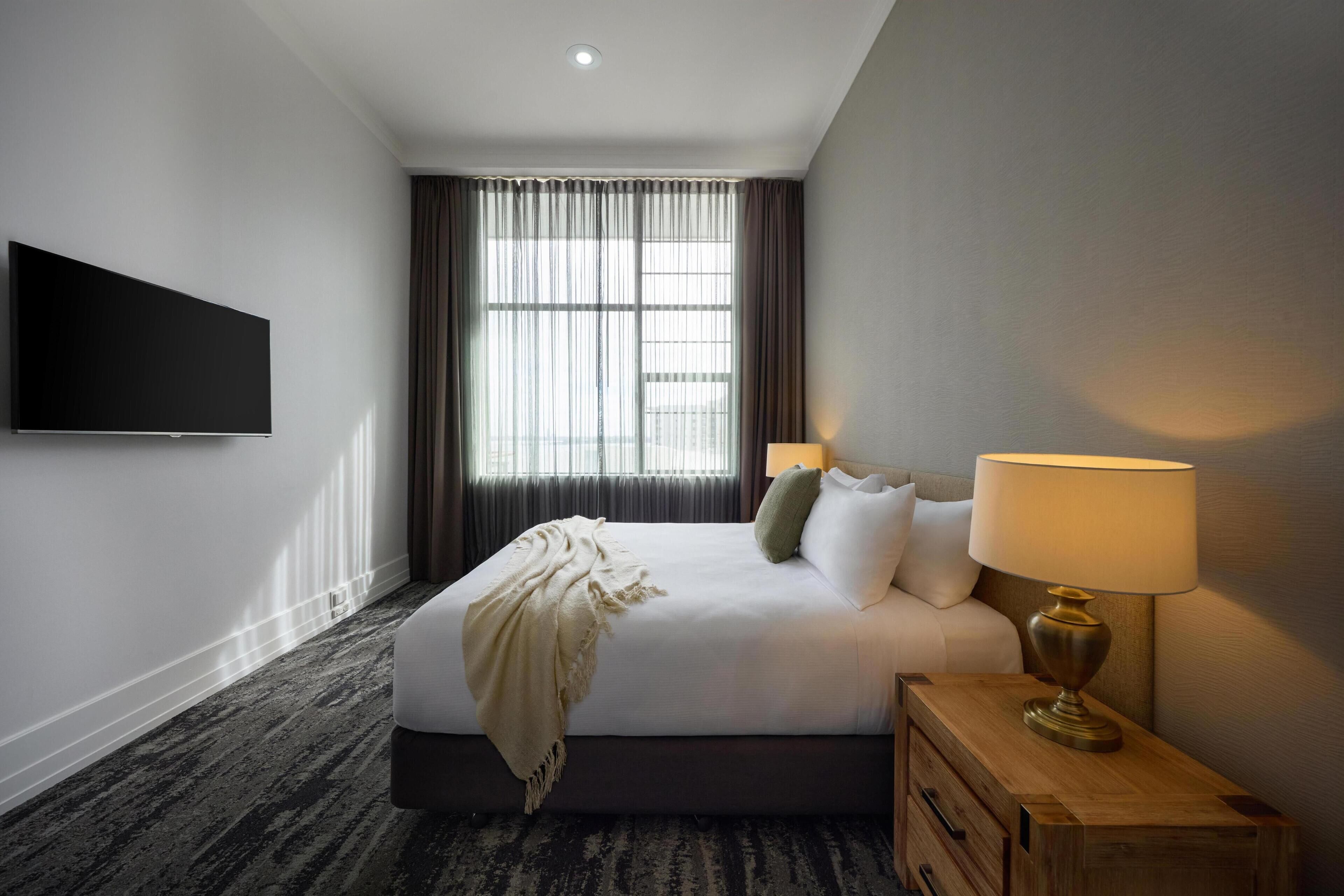 A hotel room with a bed, a TV, a lamp, a window with curtains, and a carpeted floor.