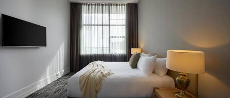 A hotel room with a bed, a TV, a lamp, a window with curtains, and a carpeted floor.