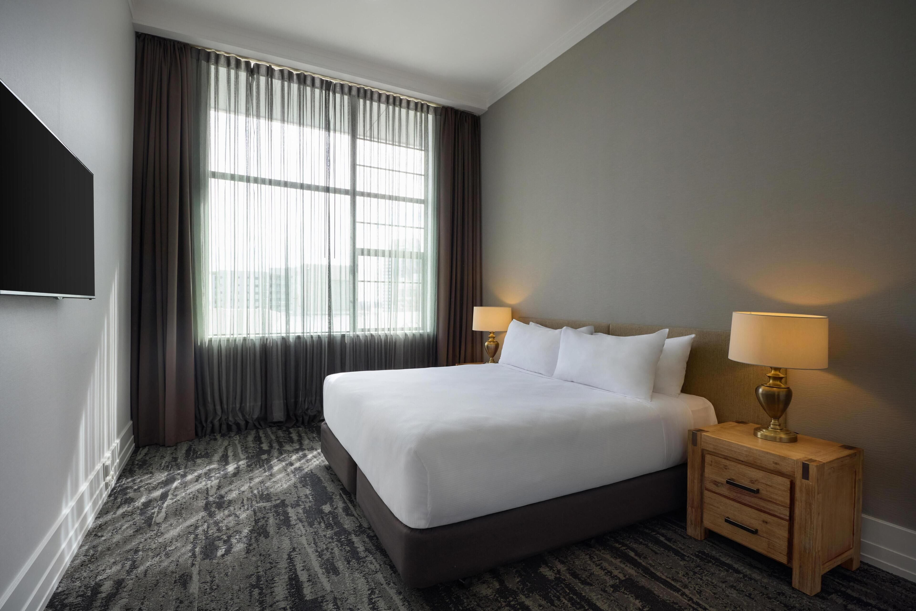 Une chambre d’hôtel avec un grand lit, deux tables de chevet avec des lampes, un téléviseur à écran plat et une fenêtre avec des rideaux.