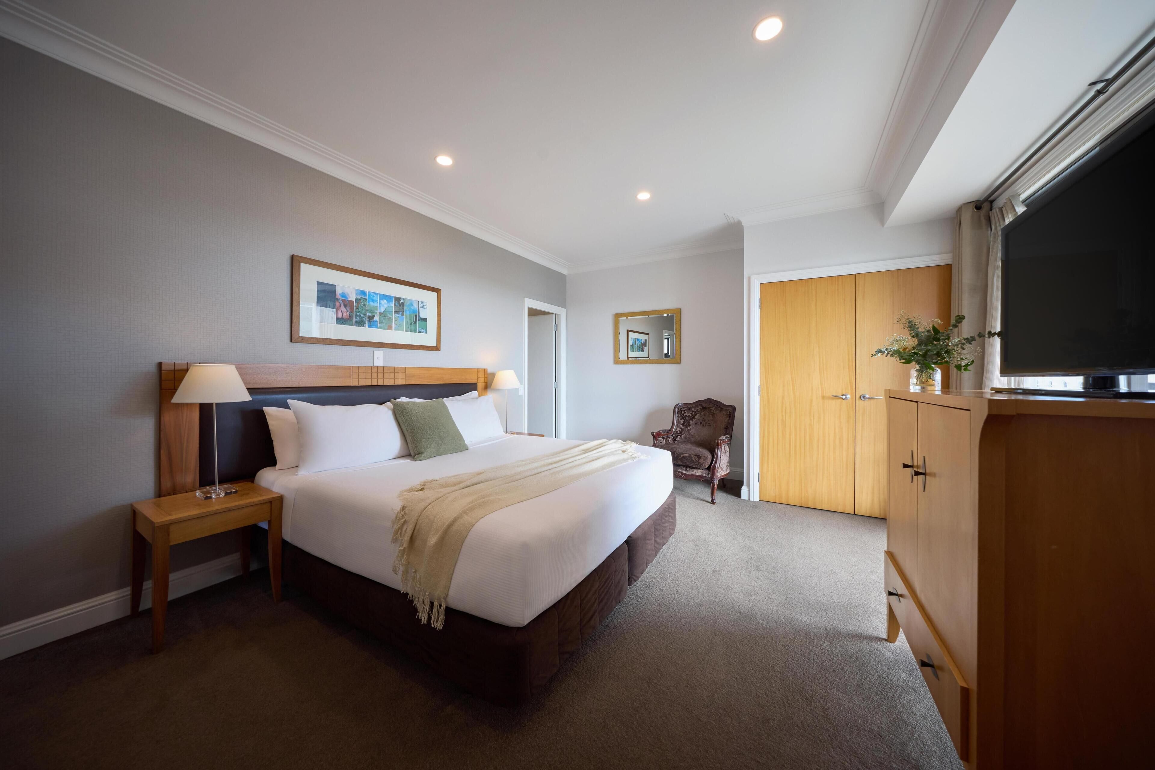 Ein Hotelzimmer mit Bett, Nachttischen, Fernseher, Kleiderschrank und einer Tür zu einem weiteren Raum.