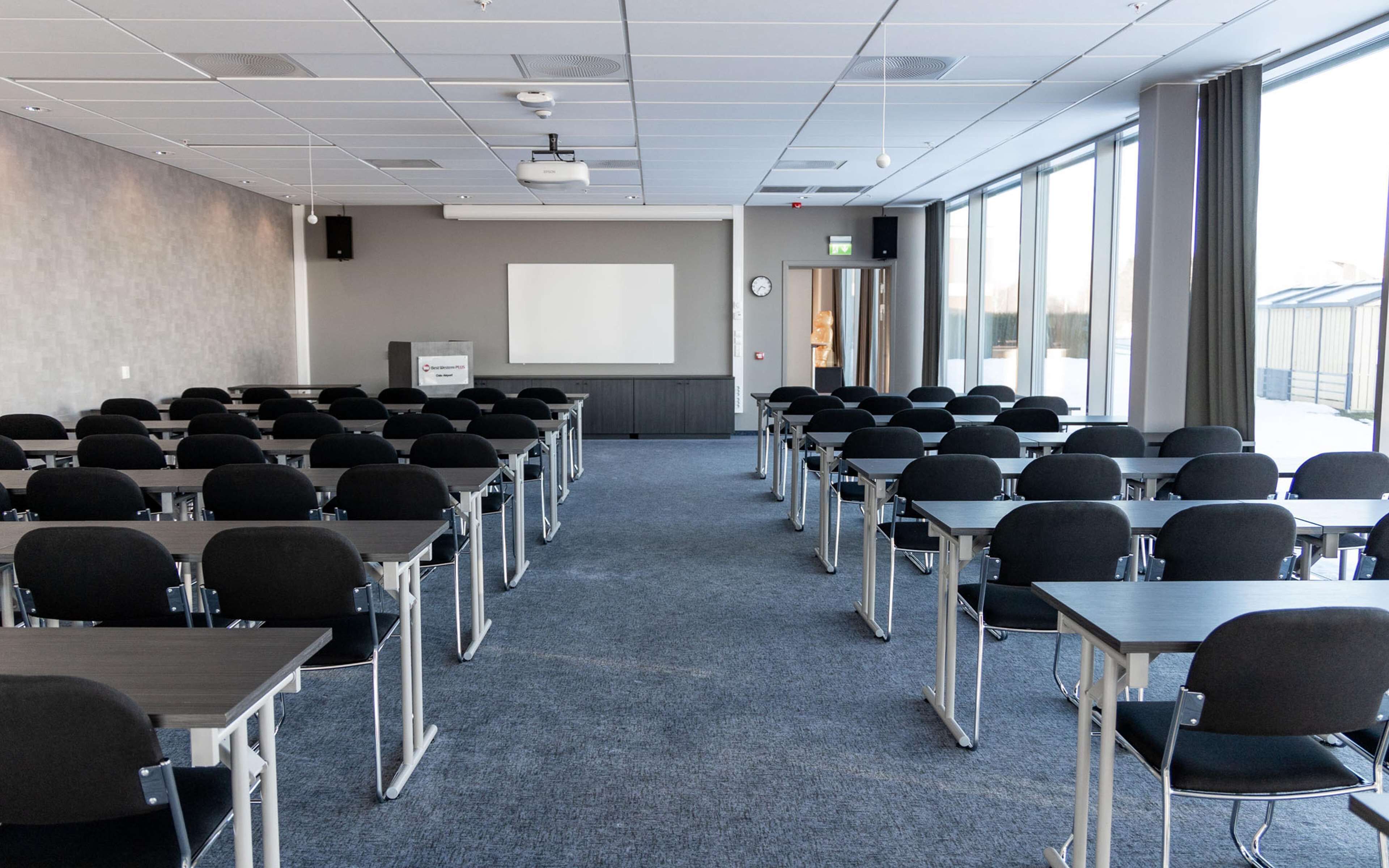 Une salle de conférence avec des rangées de tables et de chaises, un écran de projection et une grande fenêtre laissant entrer la lumière naturelle.