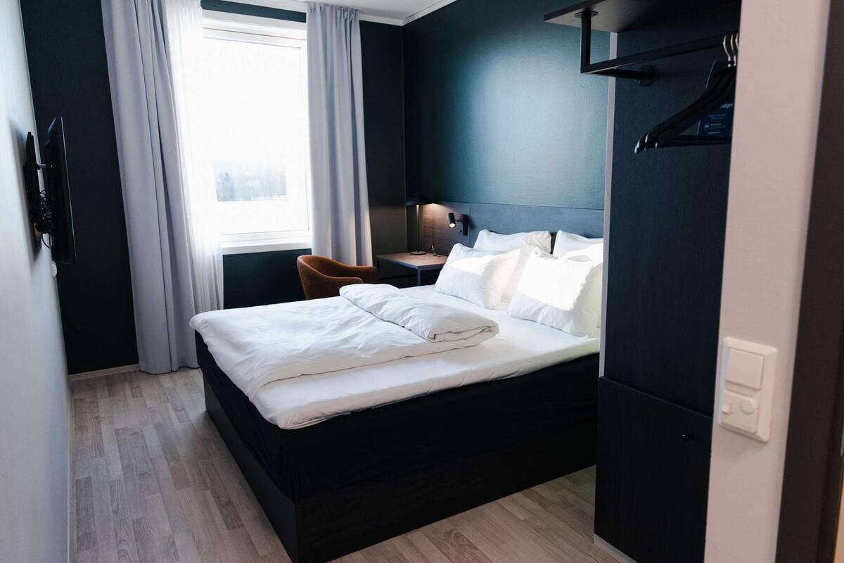 Een moderne hotelkamer met een donker bed, een houten vloer, een bureau met een lamp en uitzicht op het buitengebied door een raam met gordijnen.