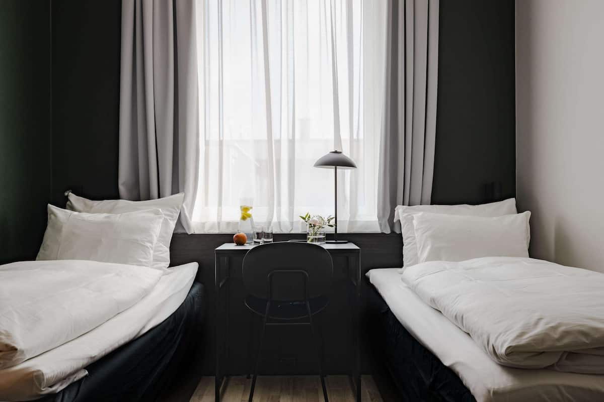 Een hotelkamer met twee eenpersoonsbedden, een bureau met stoel, een lamp en een raam met dunne gordijnen.