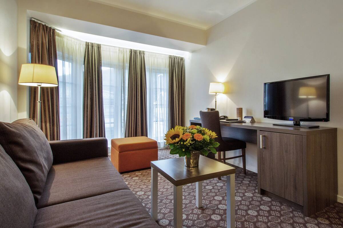 A hotel room with a sofa, a small table with a vase of flowers, a desk with a chair, a television, and curtains.