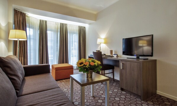 Habitación de hotel con sofá, una mesita con un florero, escritorio con silla, televisor y cortinas.