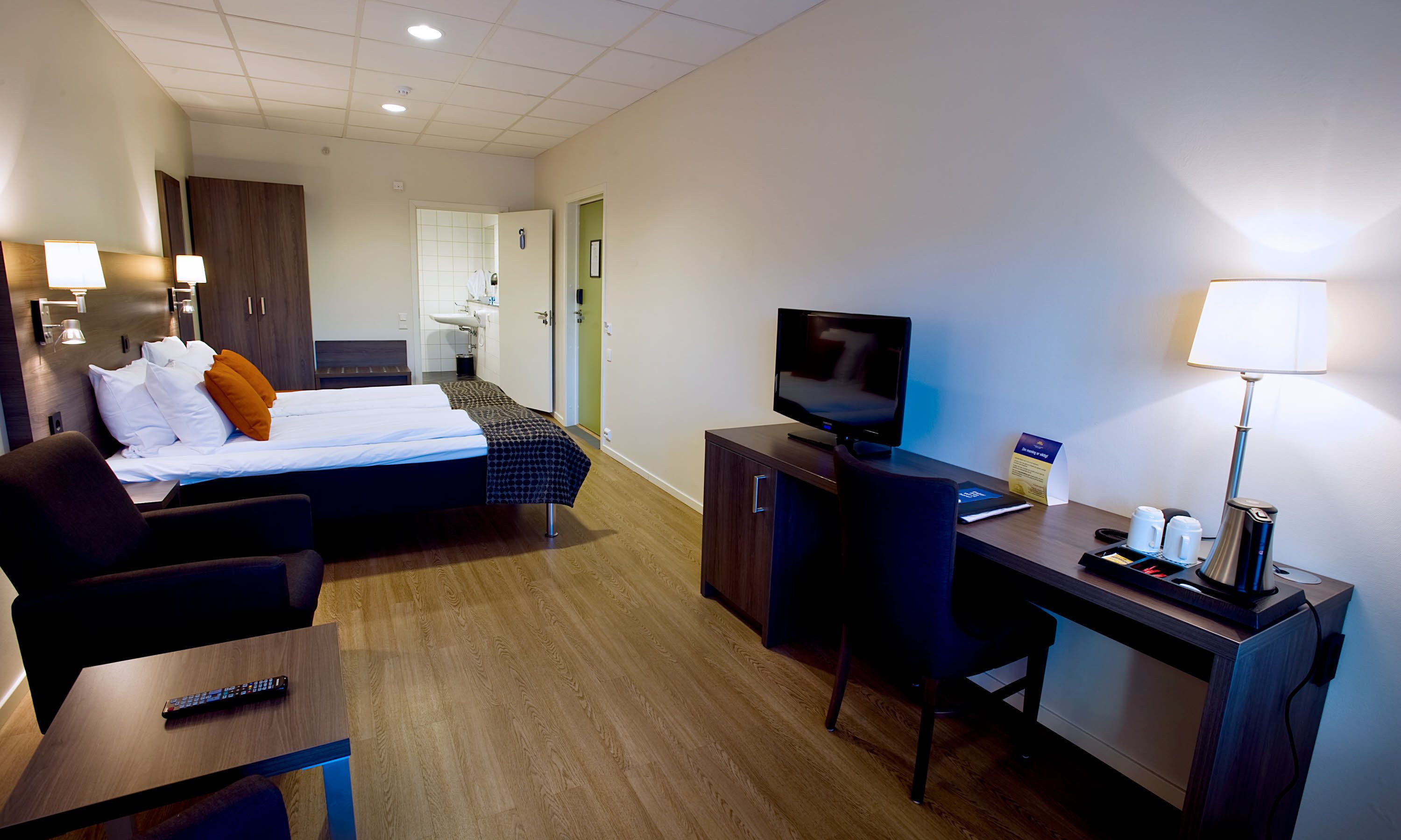 A hotel room with a bed, a desk with a TV, a chair, a remote control, and a lamp.