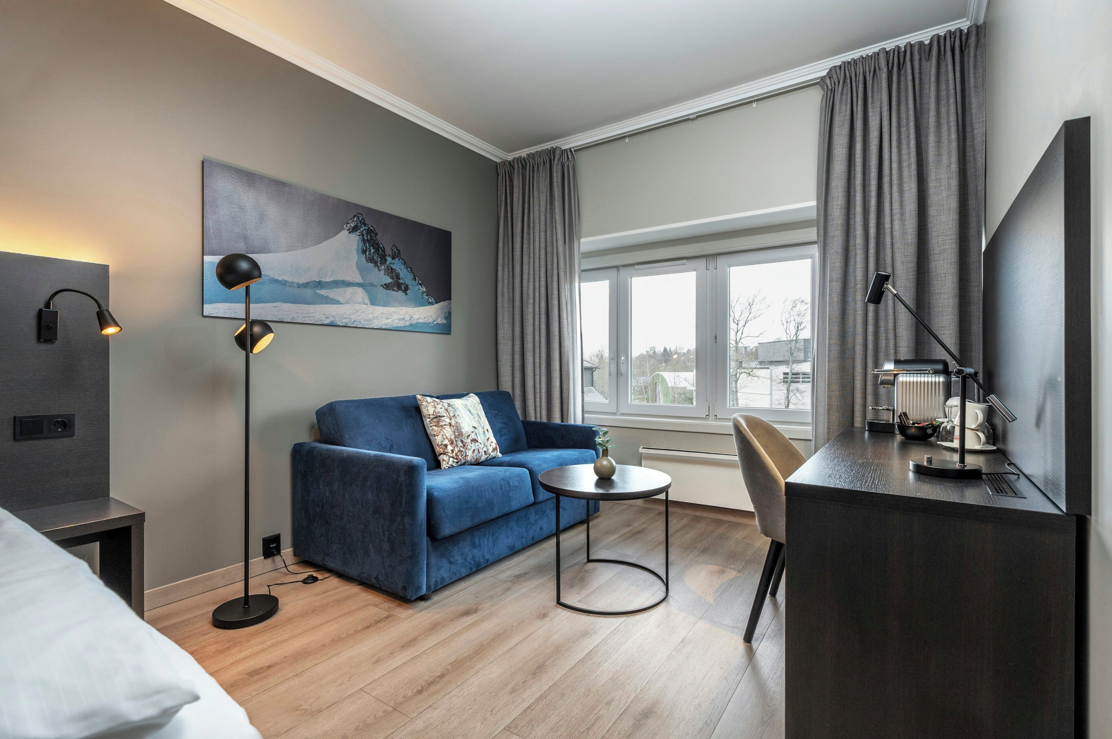 A modern hotel room with a blue sofa, a small round table, a desk with a lamp, and a large window with curtains.