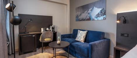 Habitación de hotel moderna con un sofá azul, una pequeña mesa redonda, un escritorio con una silla, una cafetera y un cuadro de una montaña en la pared.