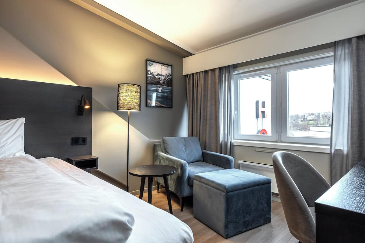 A hotel room with a bed, a desk, a chair, a small table, a lamp, and a window with curtains.