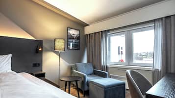 A hotel room with a bed, a desk, a chair, a small table, a lamp, and a window with curtains.