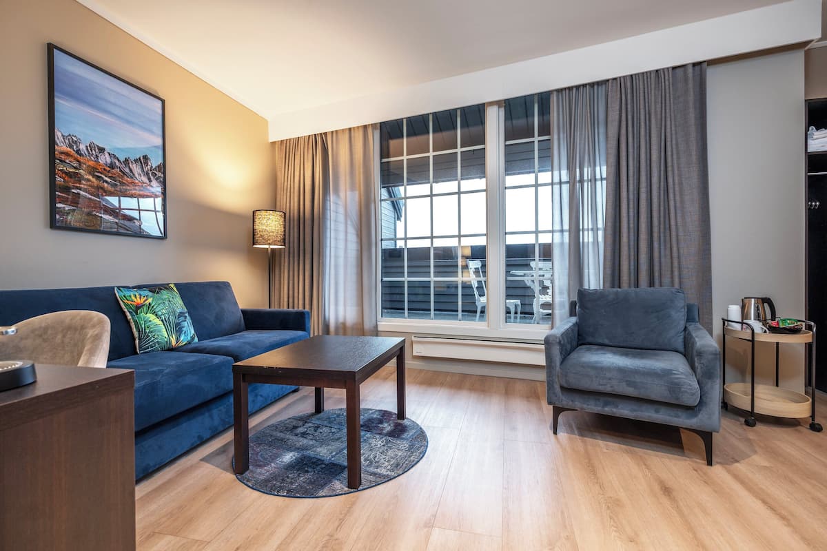 A modern living room with a blue sofa, a grey armchair, a wooden coffee table, and a large window with curtains.