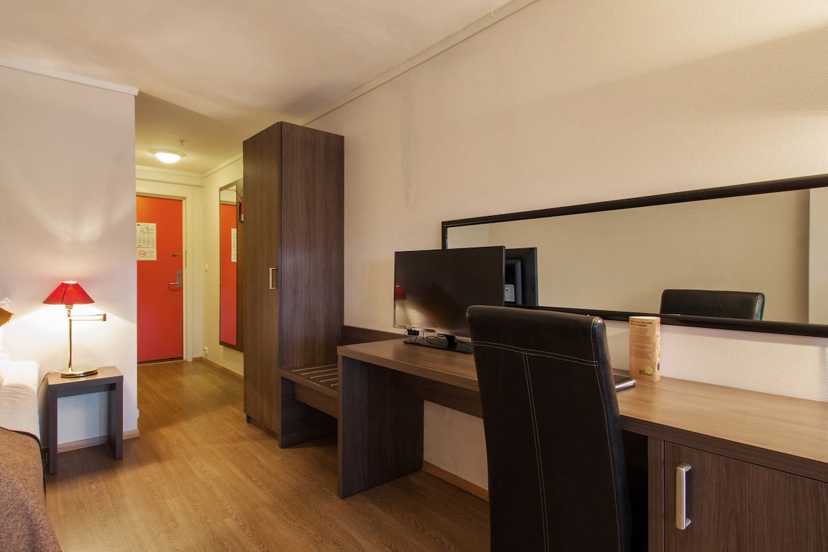 A hotel room with a wooden desk, a chair, a TV, a lamp, and a bed.