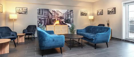 Un espace de détente moderne avec des fauteuils bleus, une table basse en bois et un grand tableau représentant un paysage urbain accroché au mur.