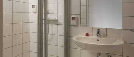A bathroom with white tiles, a glass shower enclosure, a sink with a mirror above it, and a soap dispenser on the wall.