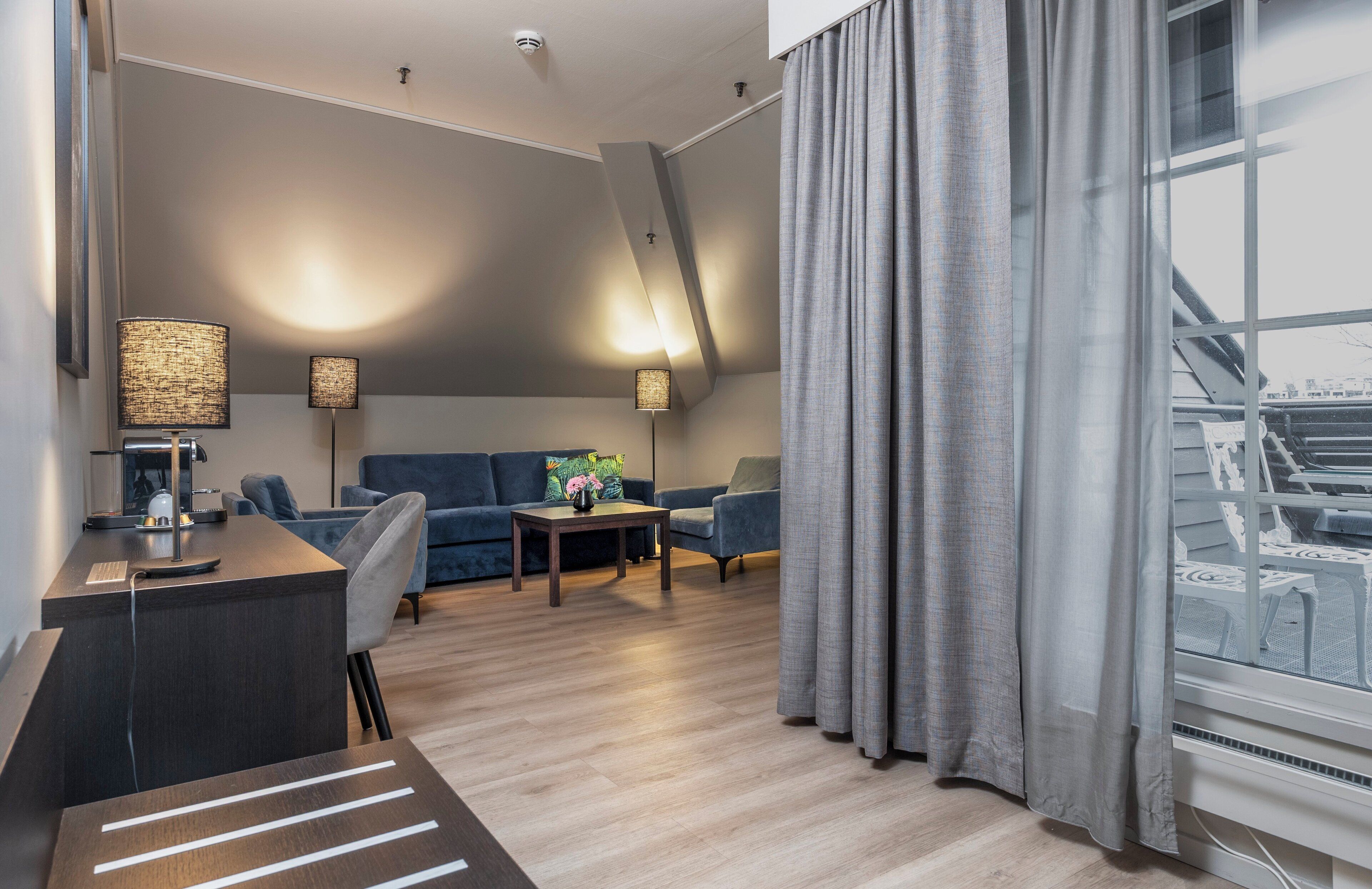 Una sala de estar moderna con un sofá, dos sillones, una mesa de centro y un escritorio con lámpara. Hay un ventanal con cortinas y vistas al exterior.
