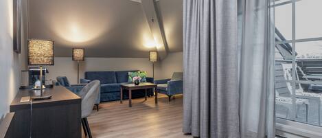 Una sala de estar moderna con un sofá, dos sillones, una mesa de centro y un escritorio con lámpara. Hay un ventanal con cortinas y vistas al exterior.