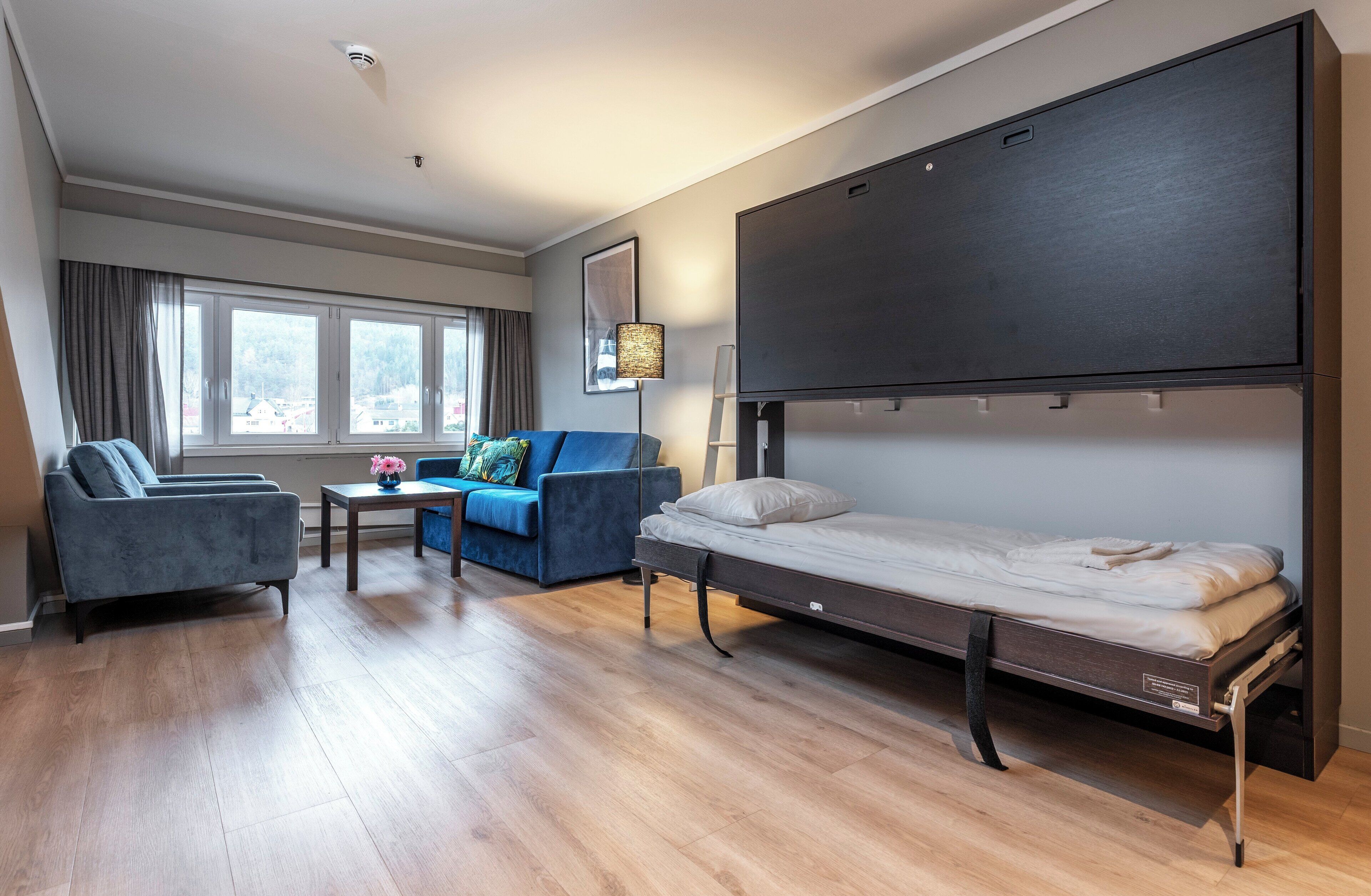 A modern hotel room with a large bed, a sofa, a small table, and a window with curtains.