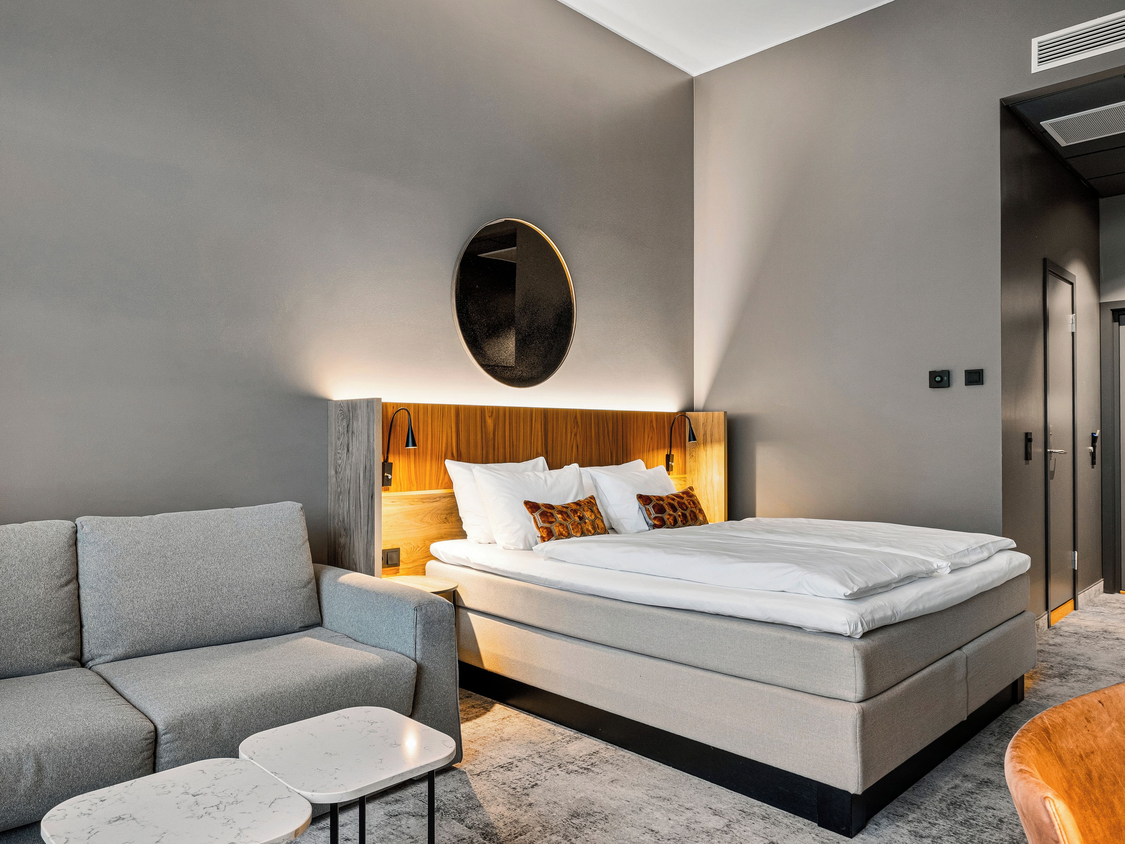 Habitación de hotel moderna con una cama grande, un sofá gris, una mesita y un cabecero de madera.