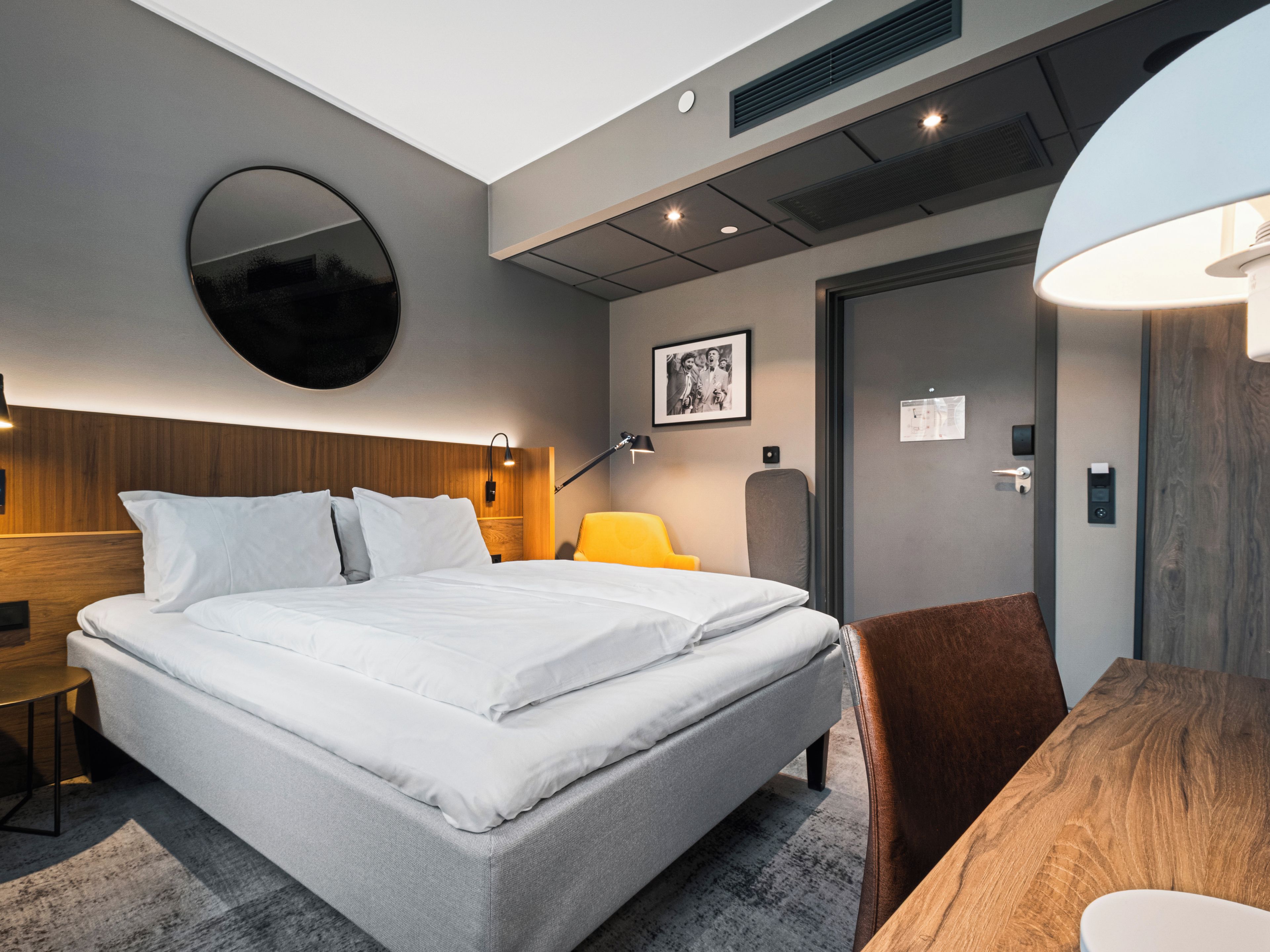 A modern hotel room with a large bed, a round mirror, a desk, and a chair.
