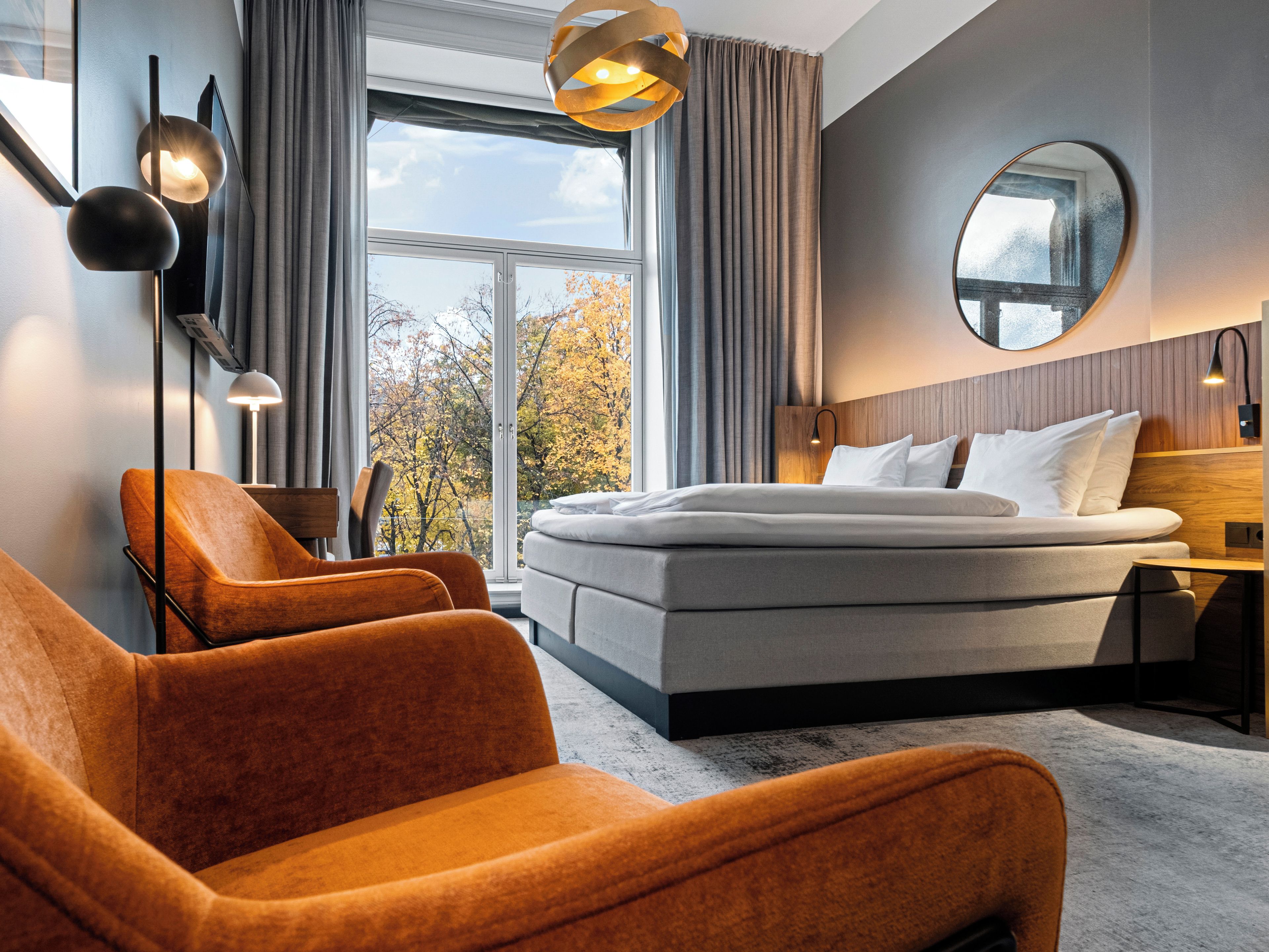 A modern hotel room with a large bed, two armchairs, a round mirror, and a view of autumn trees through the window.