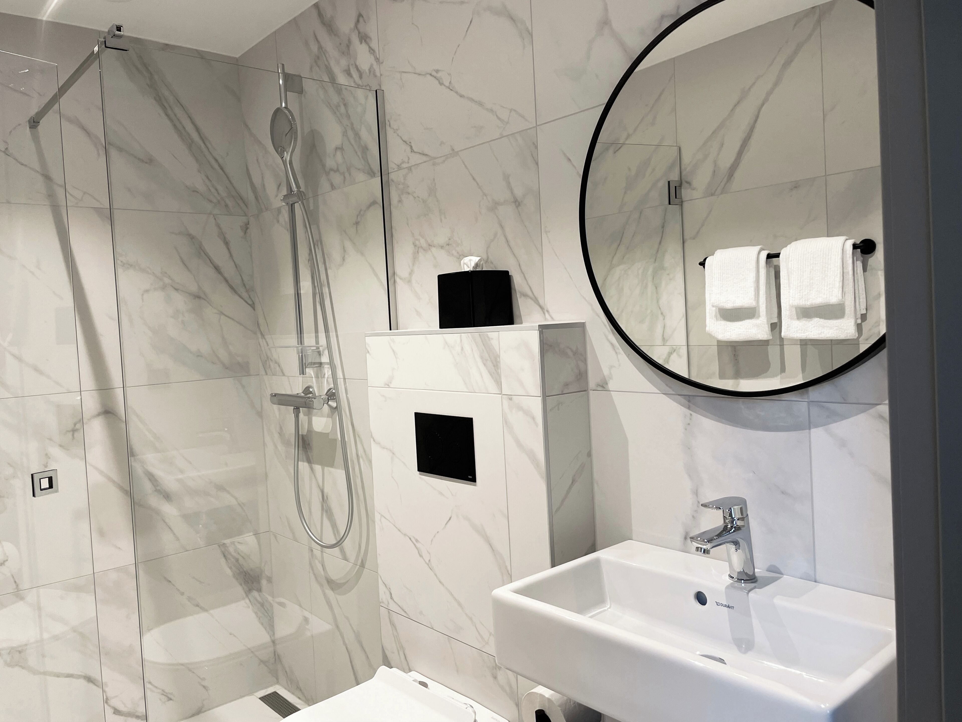A modern bathroom with a glass-enclosed shower, a white sink, and a round mirror.