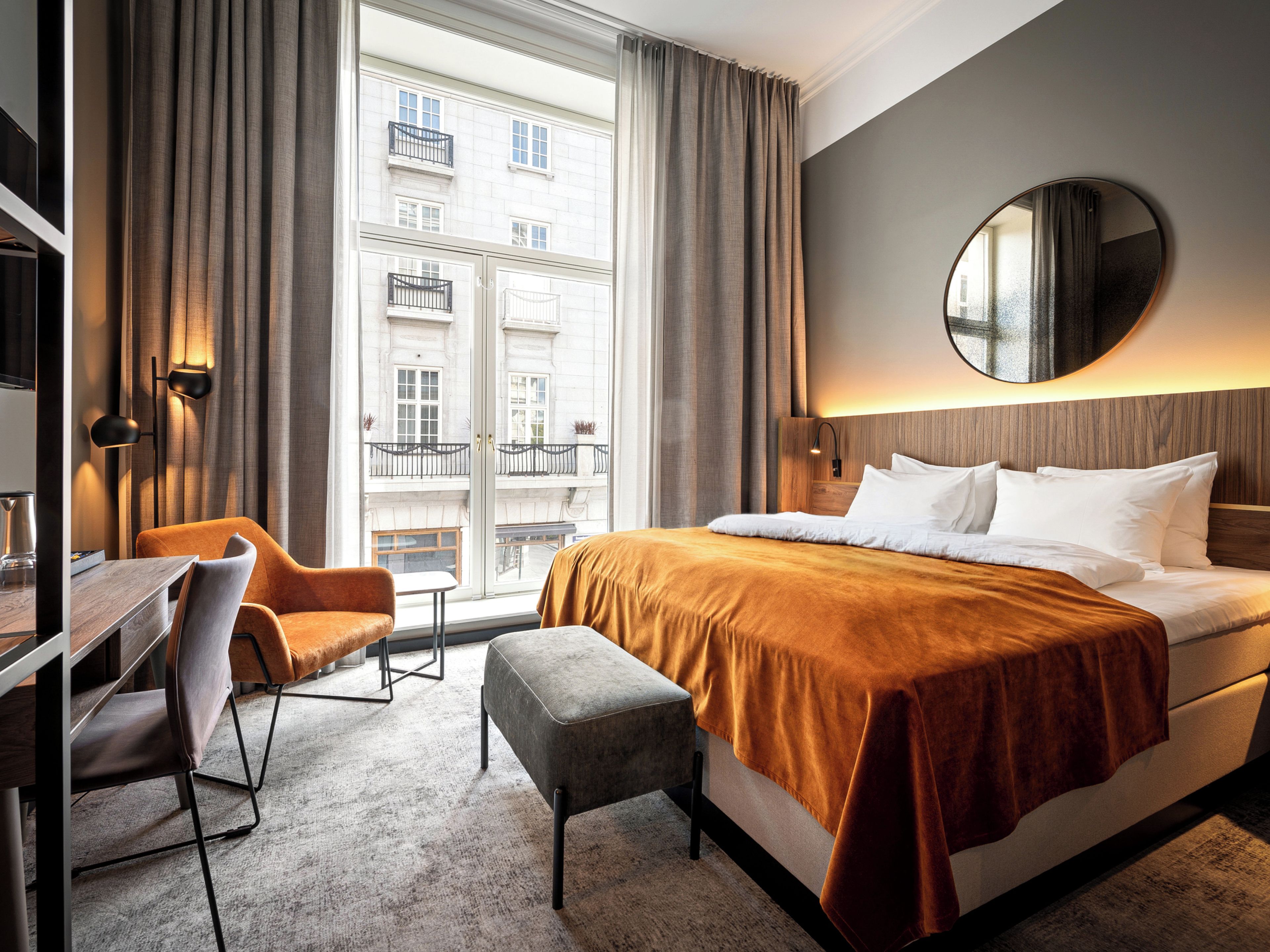 A hotel room with a large bed, a desk with a chair, a round mirror, and a view of a building through the window.