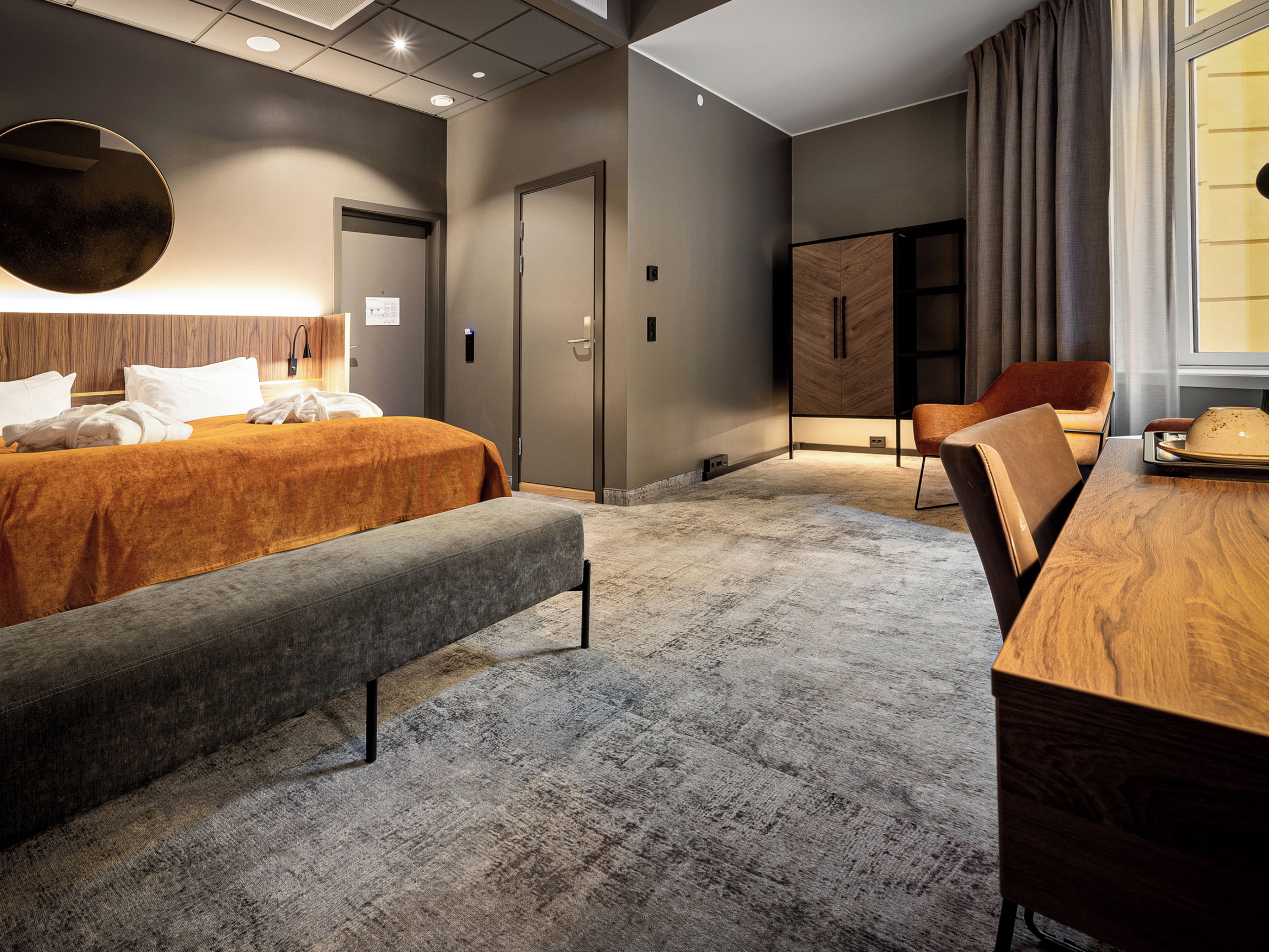 Una habitación de hotel moderna con una cama grande, un banco, un escritorio de madera y un armario.