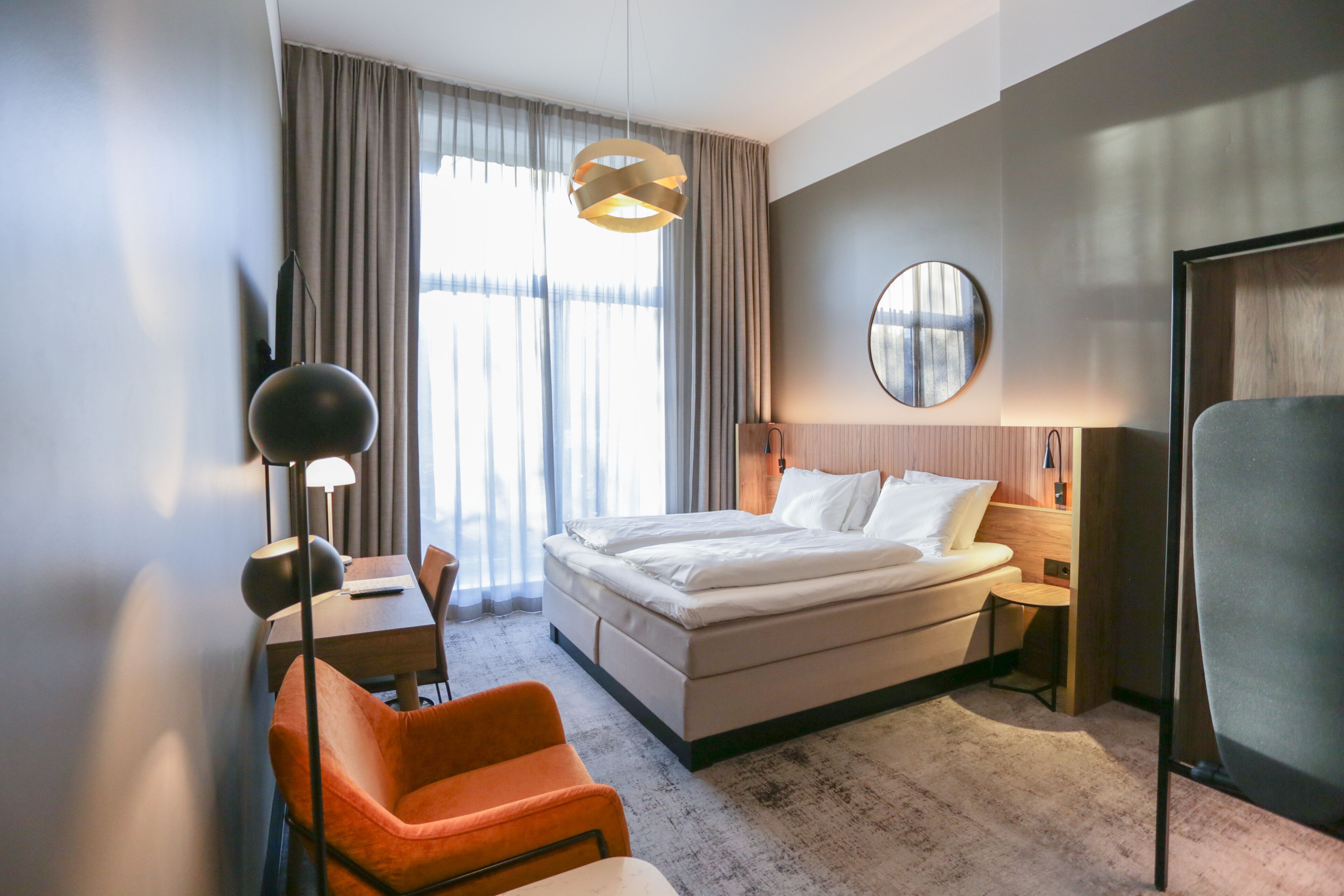 A modern hotel room with a bed, a desk, a chair, a round mirror, and a large window with curtains.