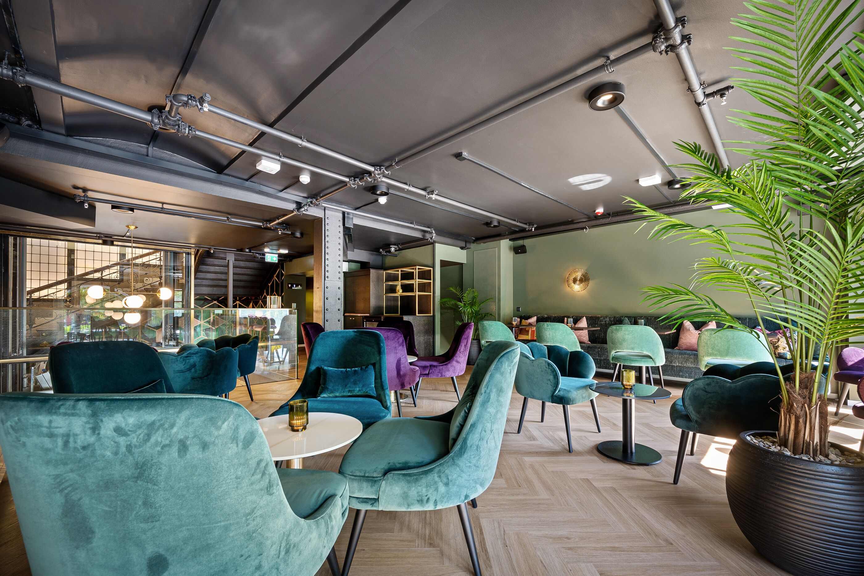 Un espace détente moderne avec des fauteuils recouverts d’un tissu turquoise et violet, une grande plante en pot et des tuyaux apparents au plafond.
