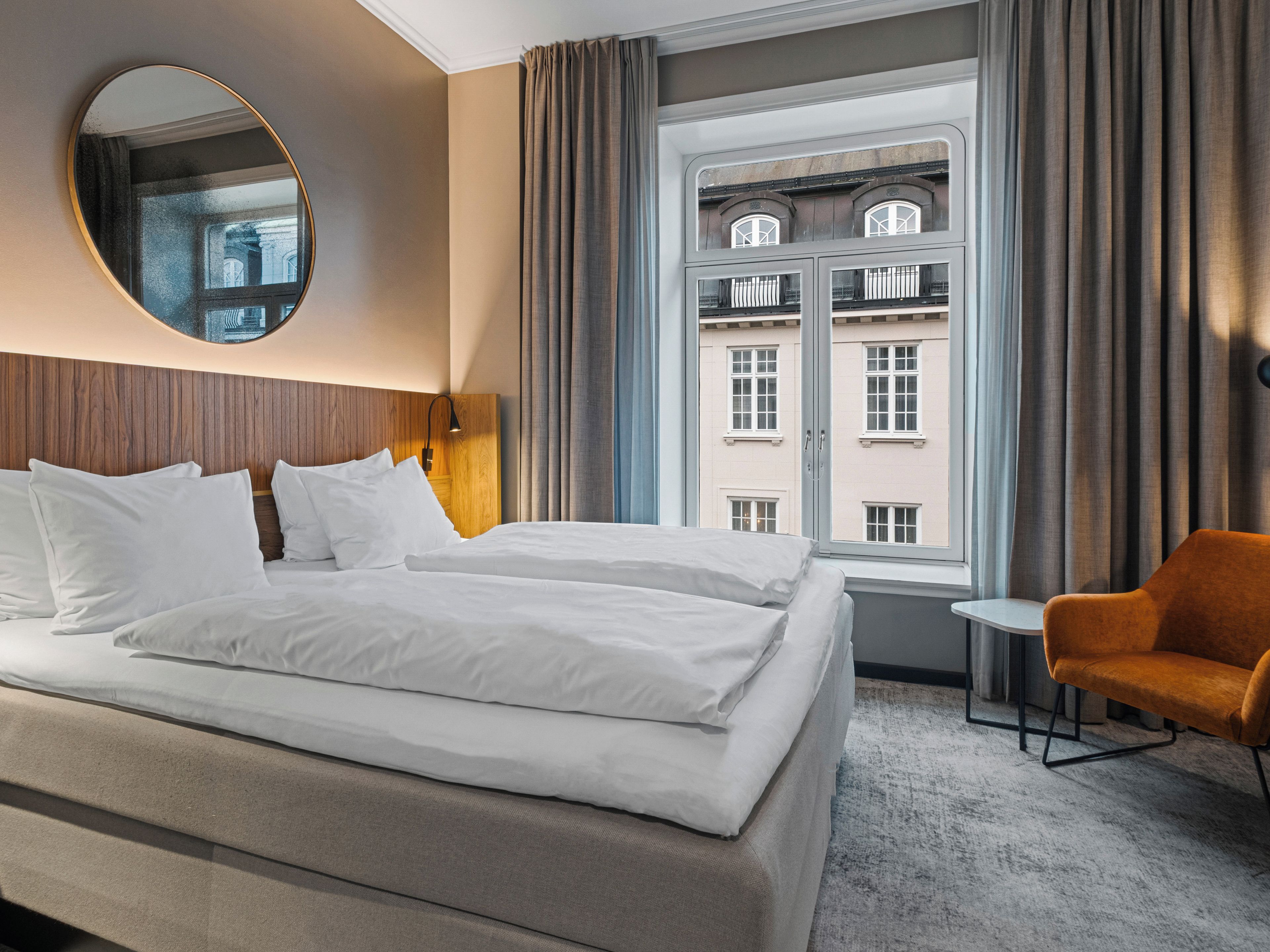 A hotel room with a bed, a chair, a round mirror, and a window with a view of buildings.