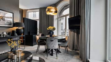 A modern hotel room with a dining area, a large window with curtains, a flat-screen TV, and a round table with chairs.