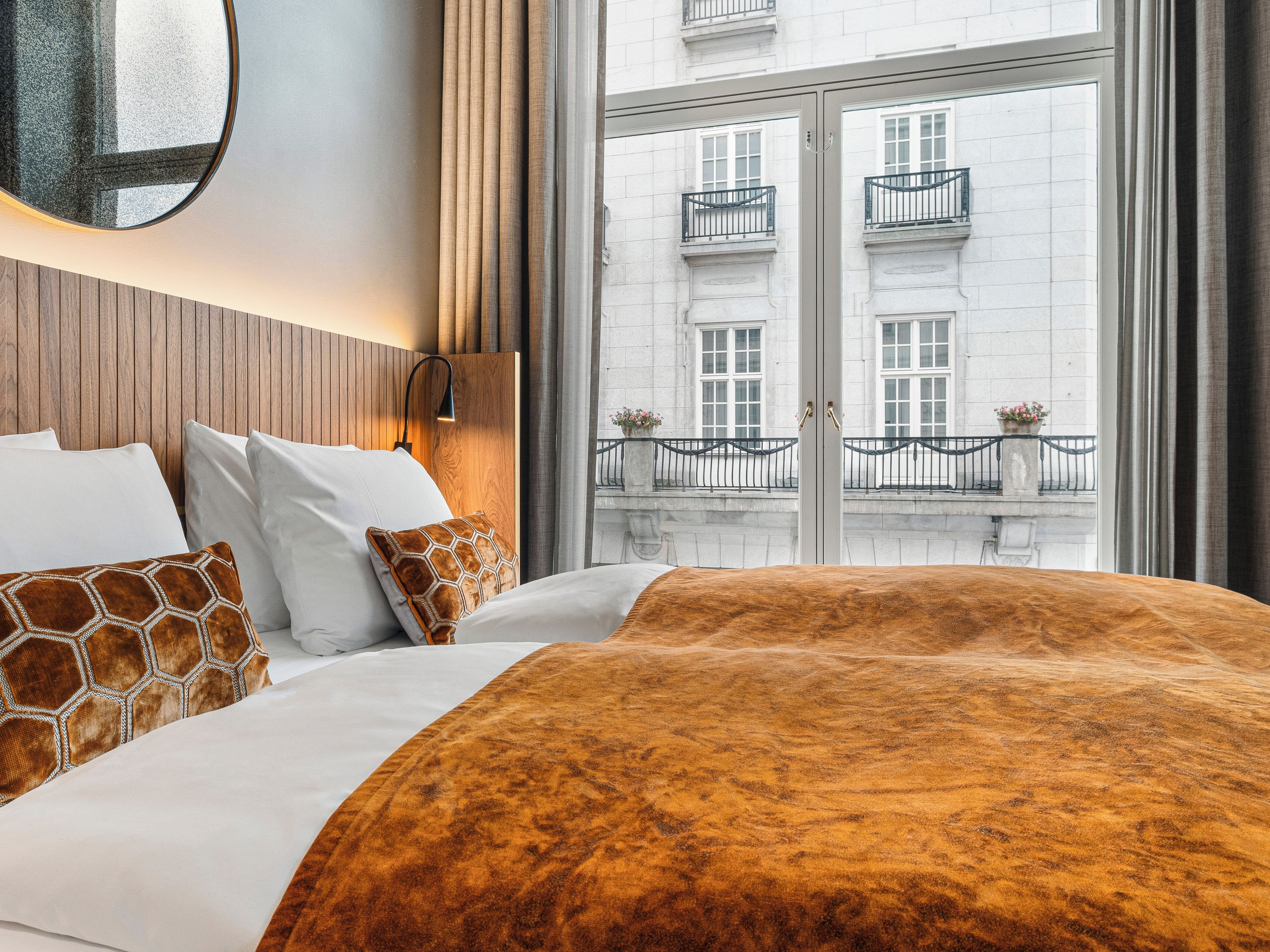 Una habitación de hotel con cama, mesita de noche, lámpara de pared y vista a un edificio con balcones.