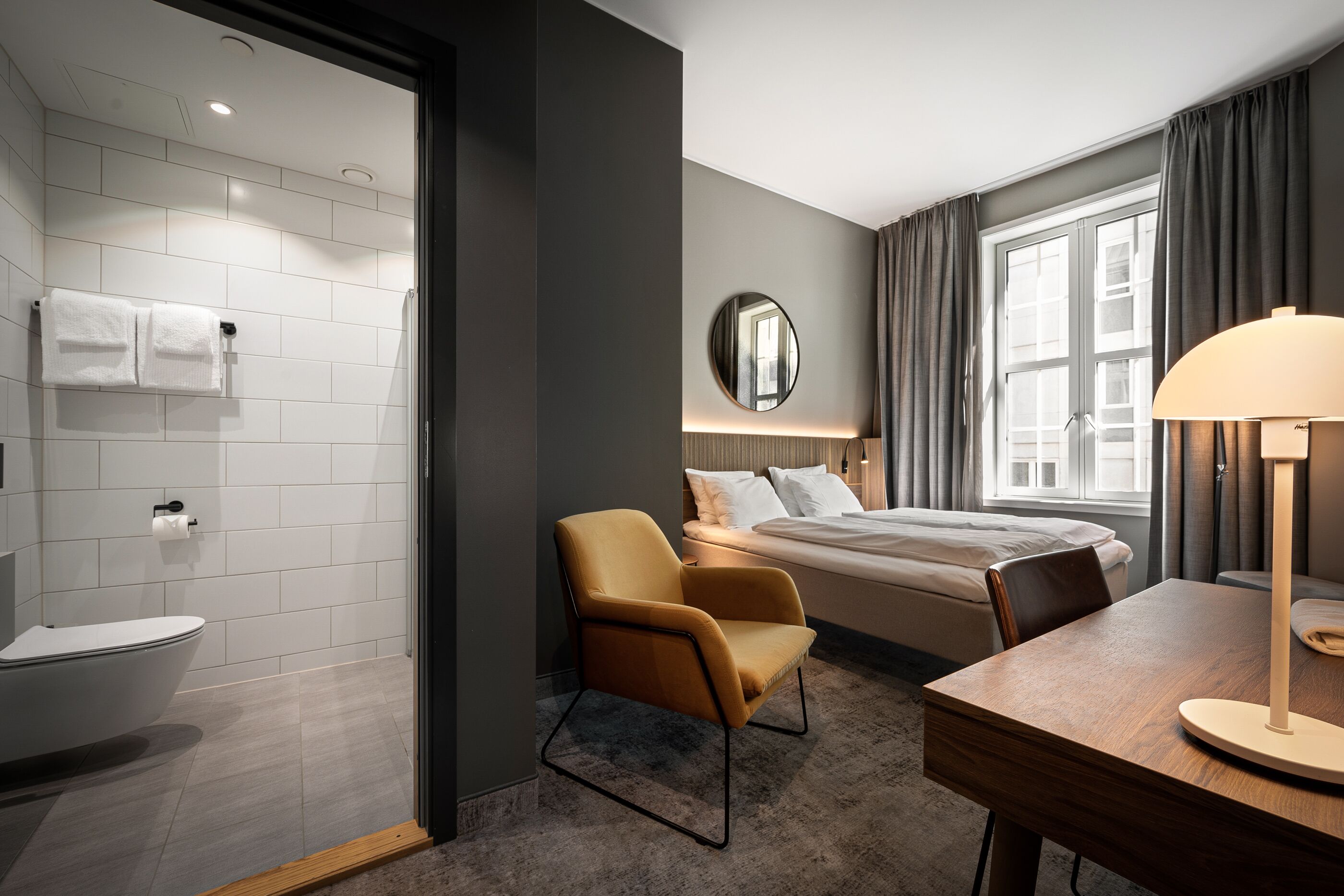 A modern hotel room with a bathroom, a bed, a chair, and a desk.