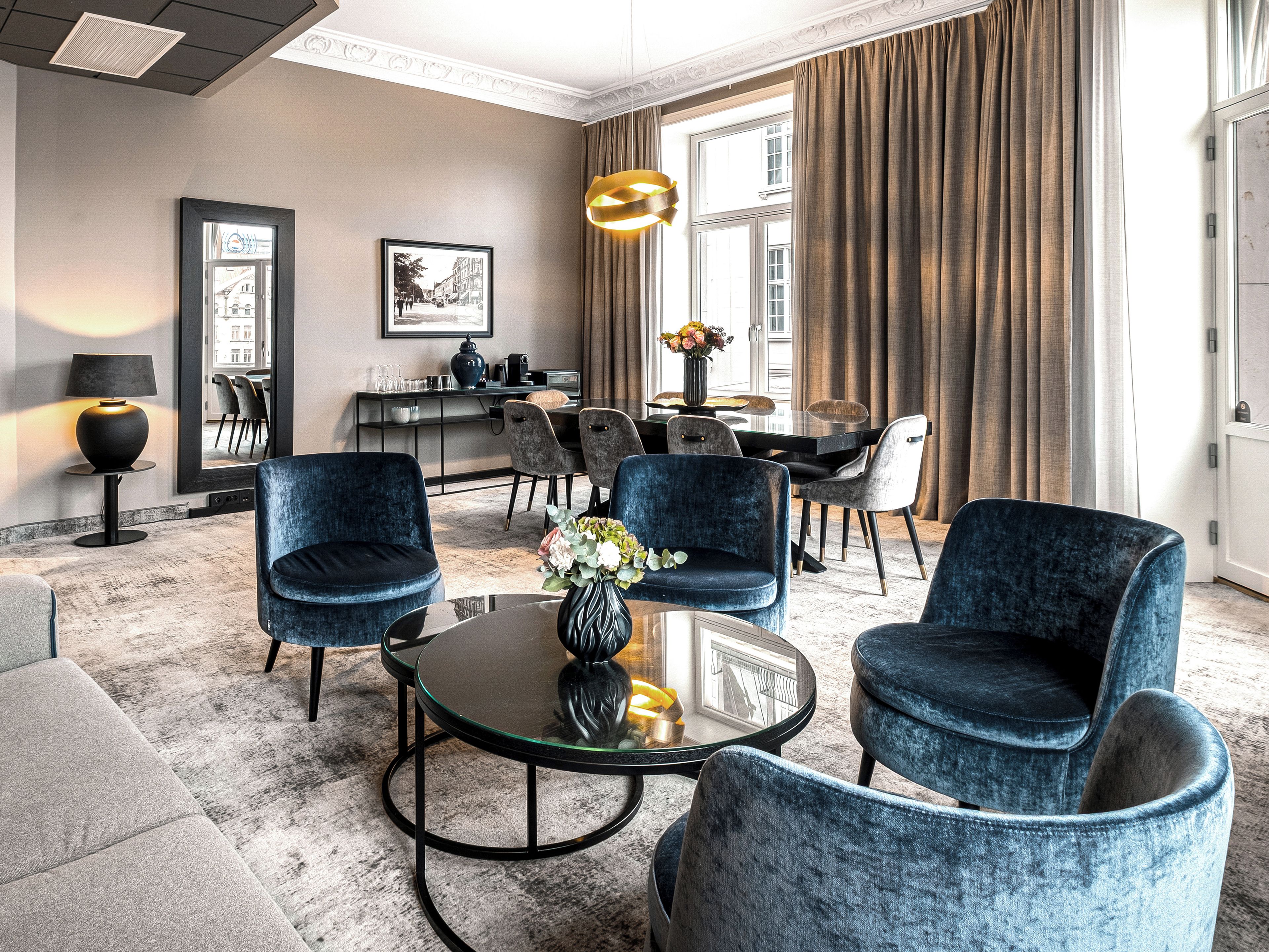 Habitación de hotel con zona de estar que incluye una mesa de centro de vidrio, dos sillones de terciopelo azul, un sofá de color oscuro, una mesita de noche con lámpara, un cuadro en la pared y un comedor con mesa y sillas.