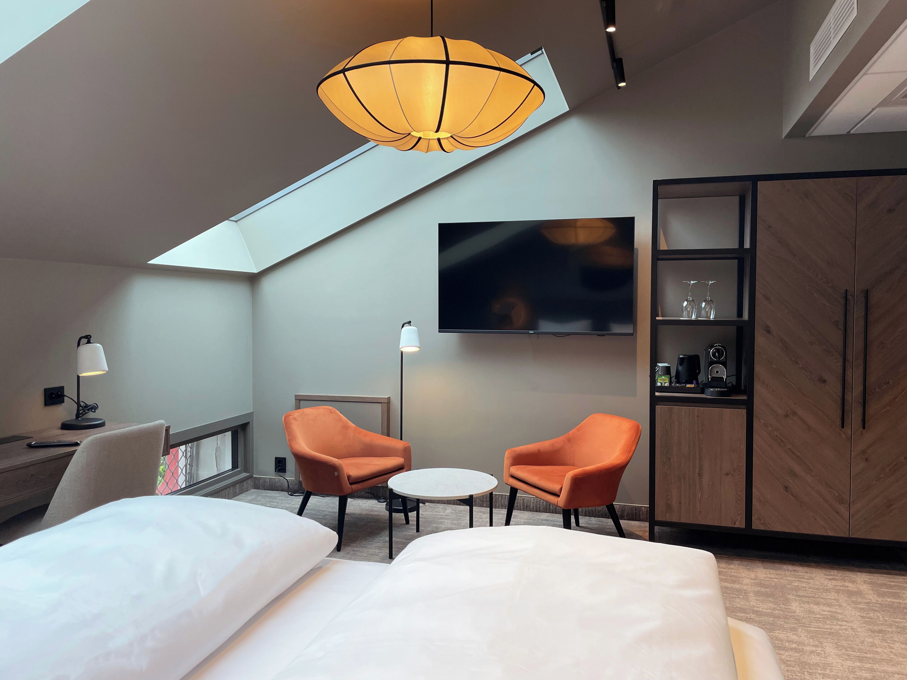 A modern hotel room with a large bed, two orange chairs, a small table, a TV mounted on the wall, and a skylight.