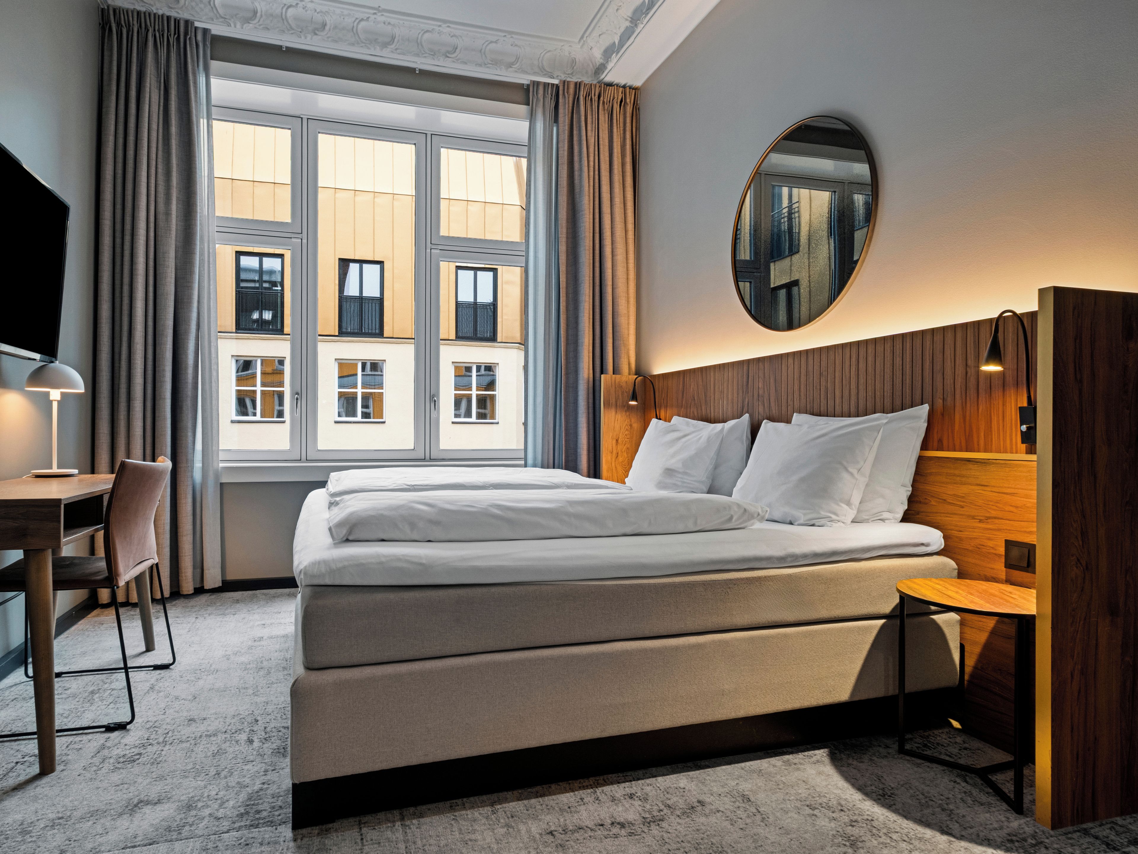 A hotel room with a large bed, a desk, a chair, a round mirror, and a view of buildings through large windows.