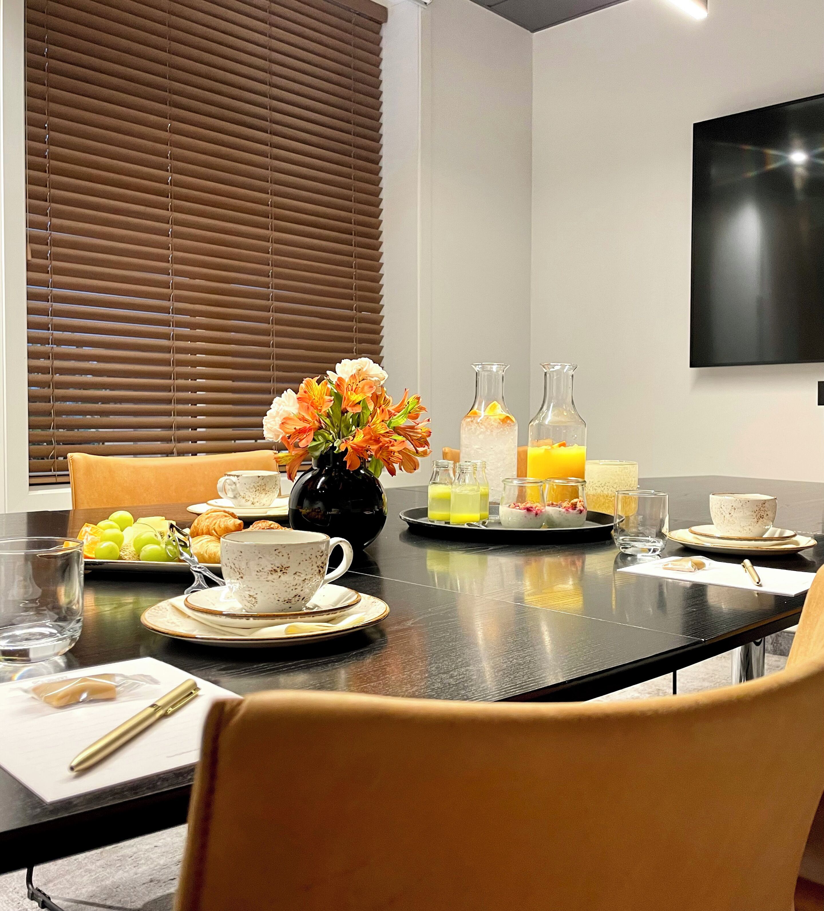 Una mesa de desayuno servida con croissants, fruta y bebidas, en una habitación moderna con televisor y persianas.